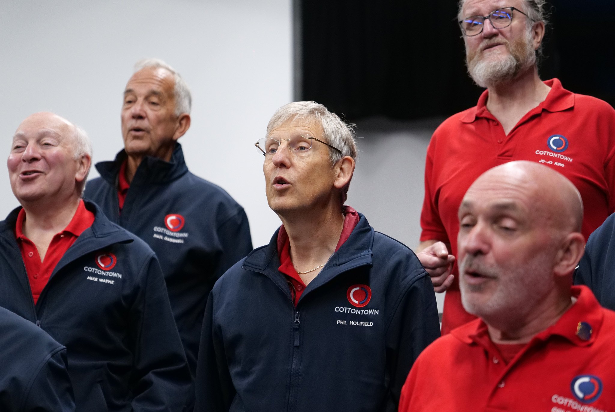 A group of six people, mostly men, standing together, wearing navy blue or red jackets with 'Cottontown' logos and name tags, appearing to sing or speak in a room with a plain background.