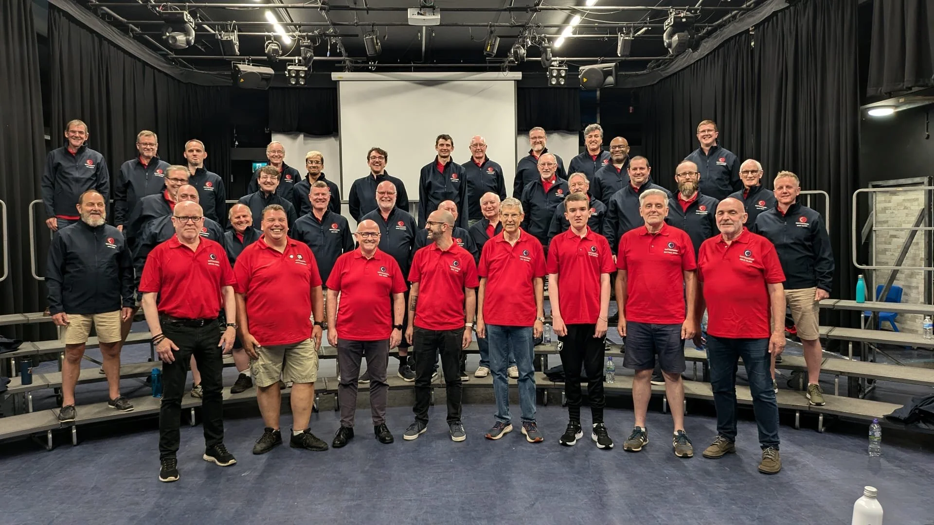 Group of men and women on a stage for a group photo, some wearing navy blue jackets and some red shirts, in a theater or auditorium setting.
