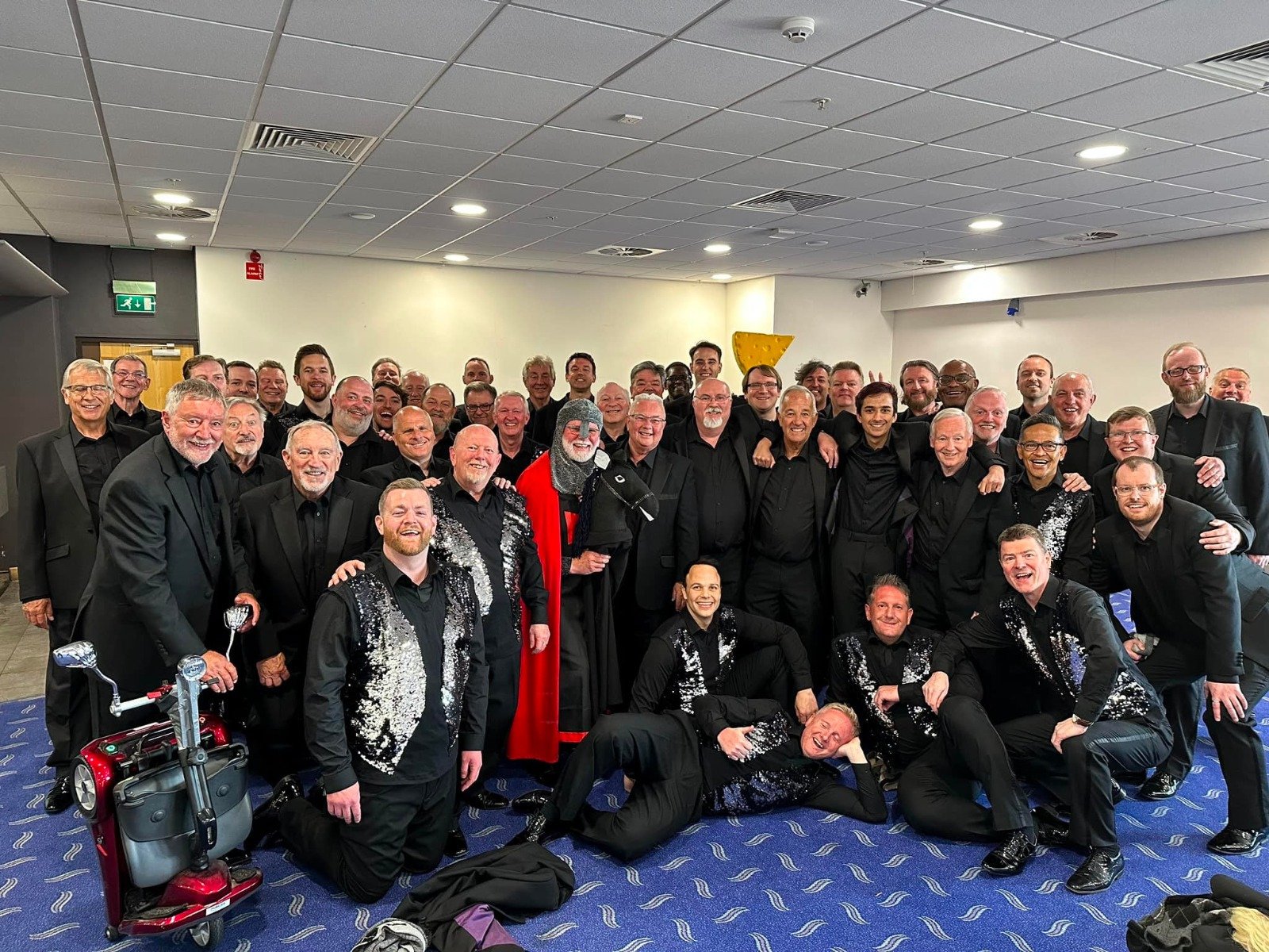 A large group of men dressed in black suits, some with glittery vests, posing for a photo in an indoor setting, with a few men in front on the floor and some wearing glasses, several smiling and making celebratory gestures.