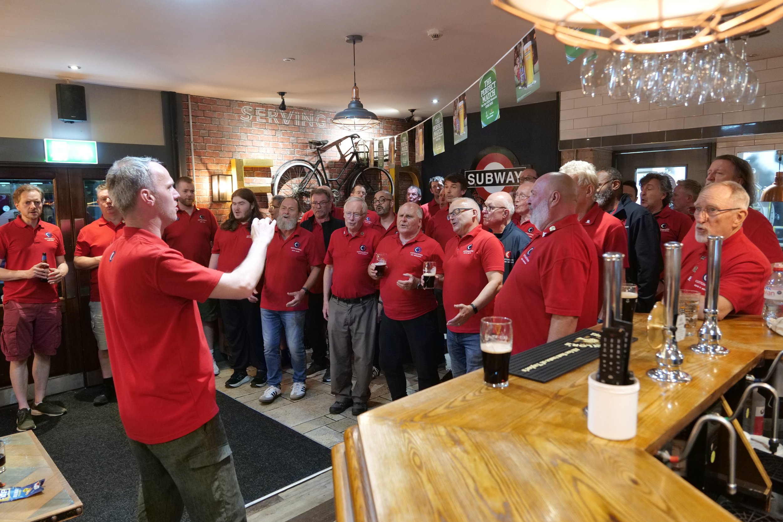 A man in a red shirt addresses a group of people, also in red shirts, gathered inside a pub or restaurant. The group appears to be listening attentively, with some holding drinks. The setting includes a wooden bar counter with taps, a bicycle mounted