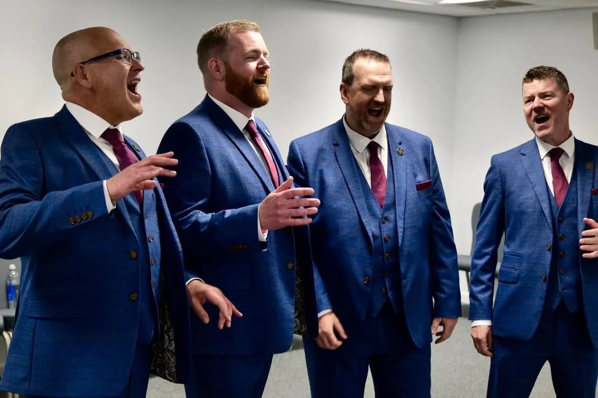 Four men in matching blue suits and maroon ties singing and laughing together in a room.