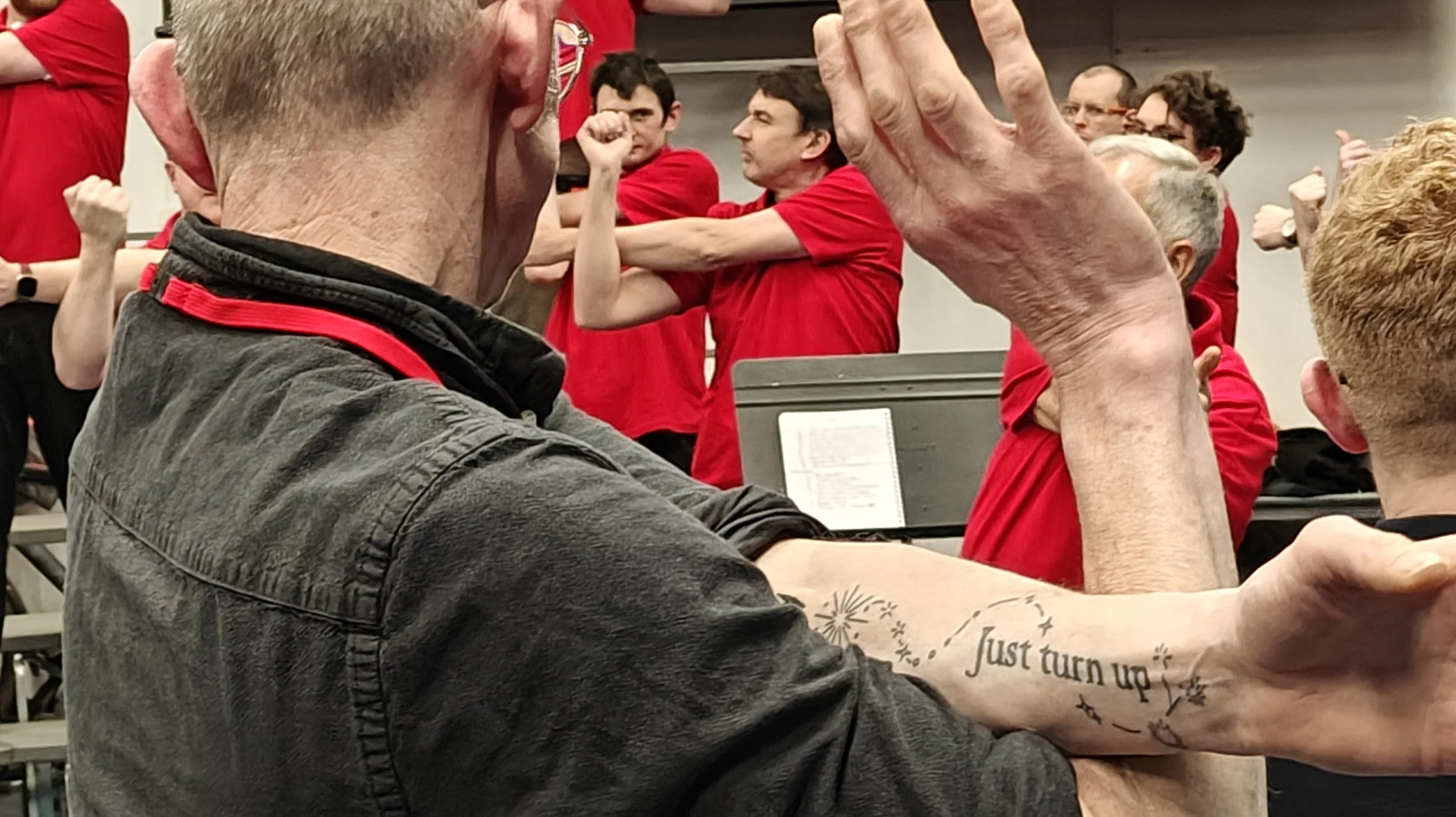 Group of people participating in a group activity, with one person in the foreground flexing his arm, showing a tattoo that reads 'Just turn up' surrounded by decorative firework-style designs.
