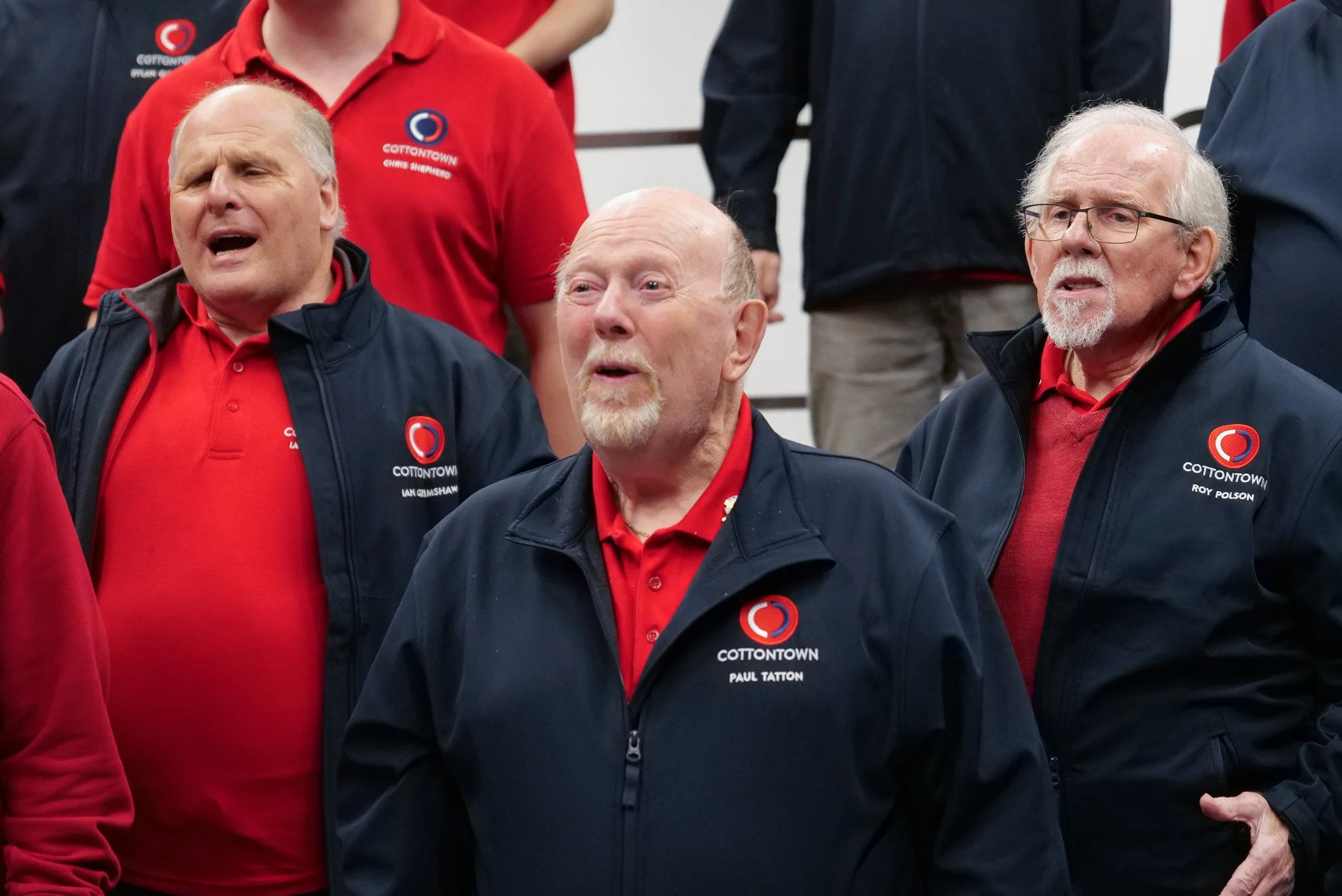 Group of men wearing red shirts and black jackets with the 'COTTONTOWN' logo, singing or shouting together.