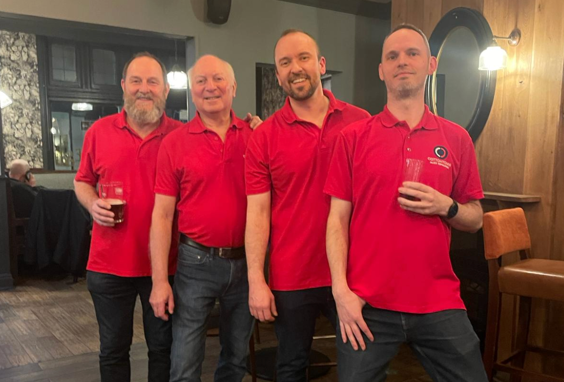 Four men standing side by side in a restaurant, wearing matching red polo shirts with a logo, and holding drinks, smiling at the camera.