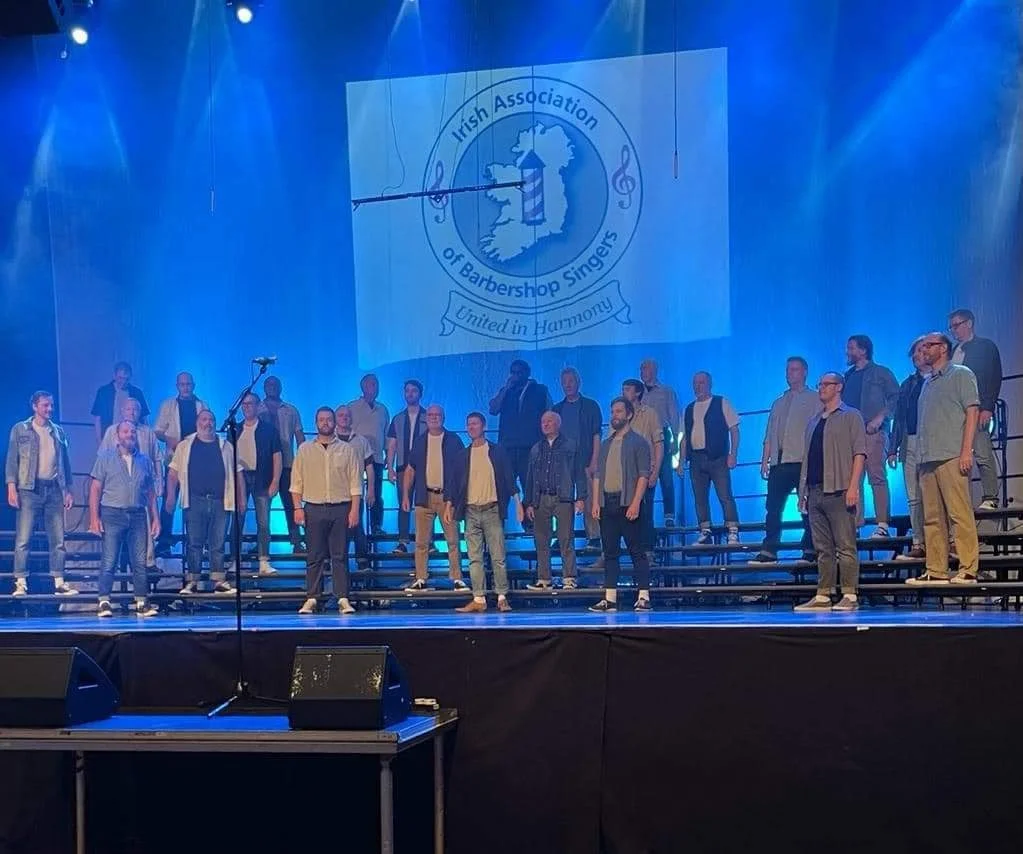 Chorus of men singing on stage with blue lighting, with a large banner in the background displaying the logo of the Irish Association of Barbershop Singers, featuring a map of Ireland and the tagline "United in Harmony."
