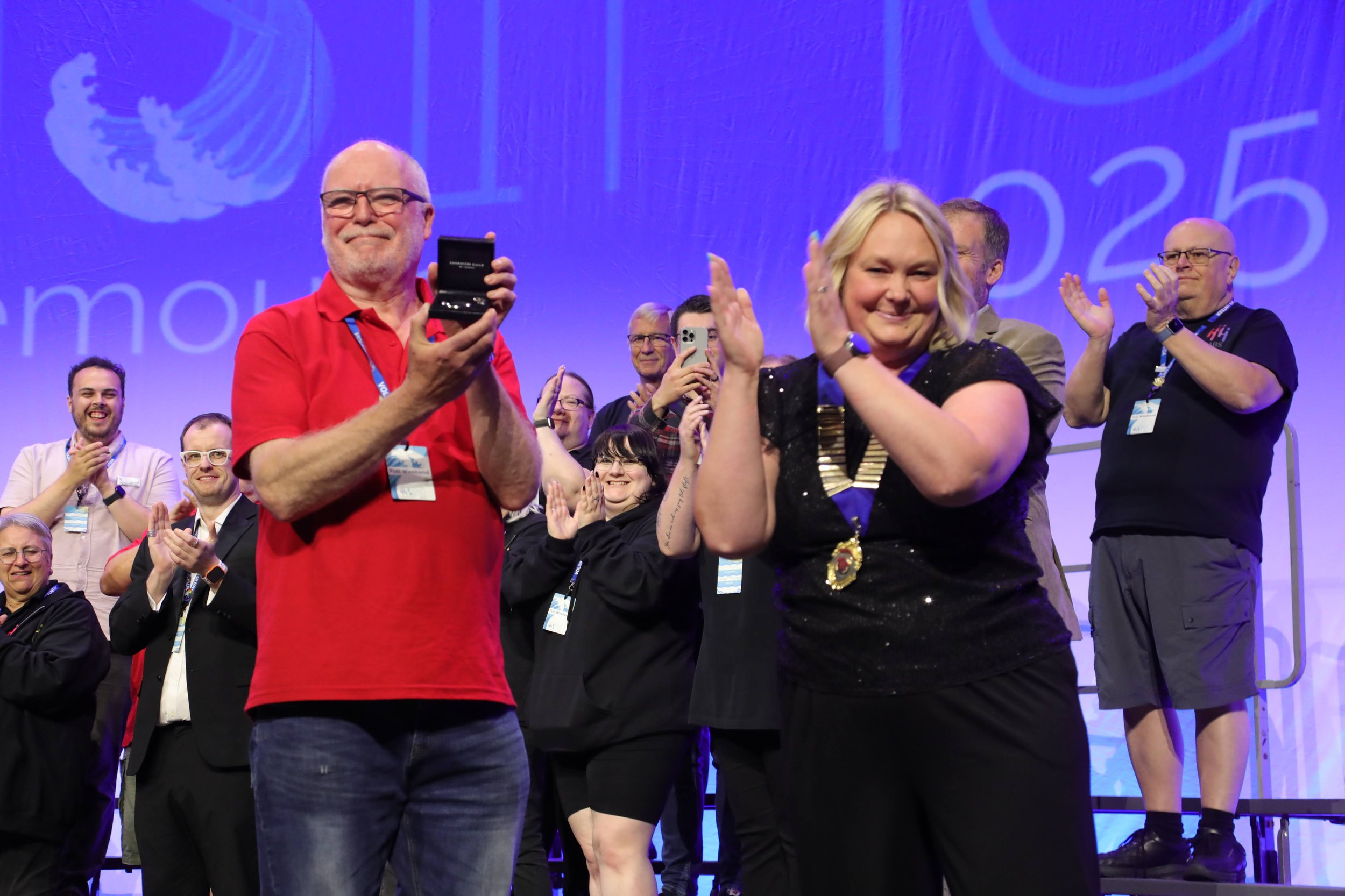 People on stage at an event, with one man in a red shirt holding an award or plaque and a woman in black with a medal around her neck, both smiling and clapping, surrounded by an audience clapping and taking photos.