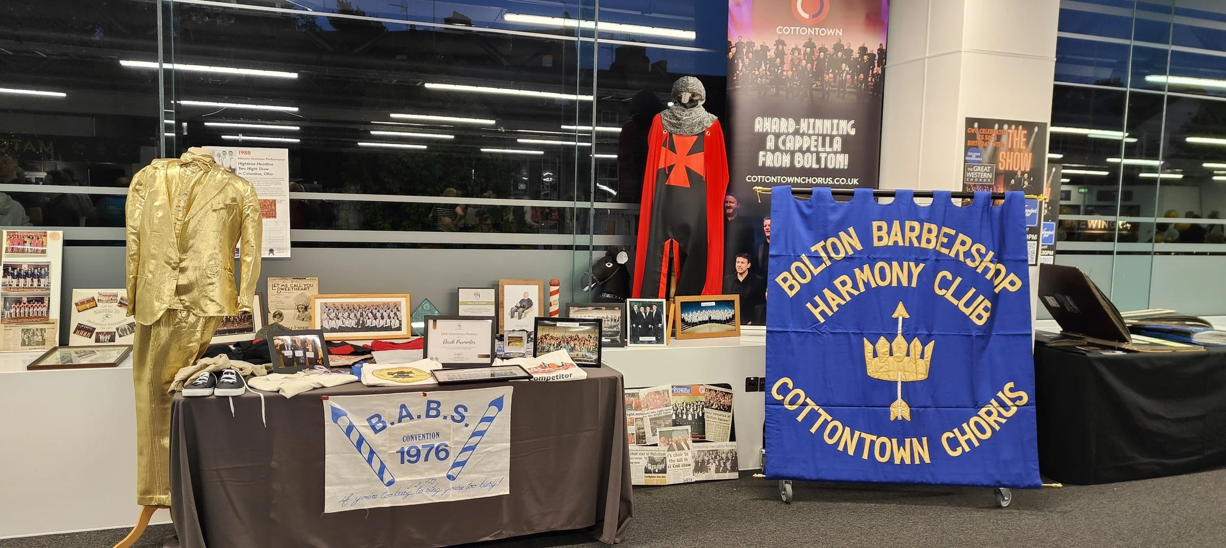 Display of vintage clothing, framed photographs, and memorabilia, including a gold suit, a cape with a cross, and a blue banner for Bolton Barbershop Harmony Club.