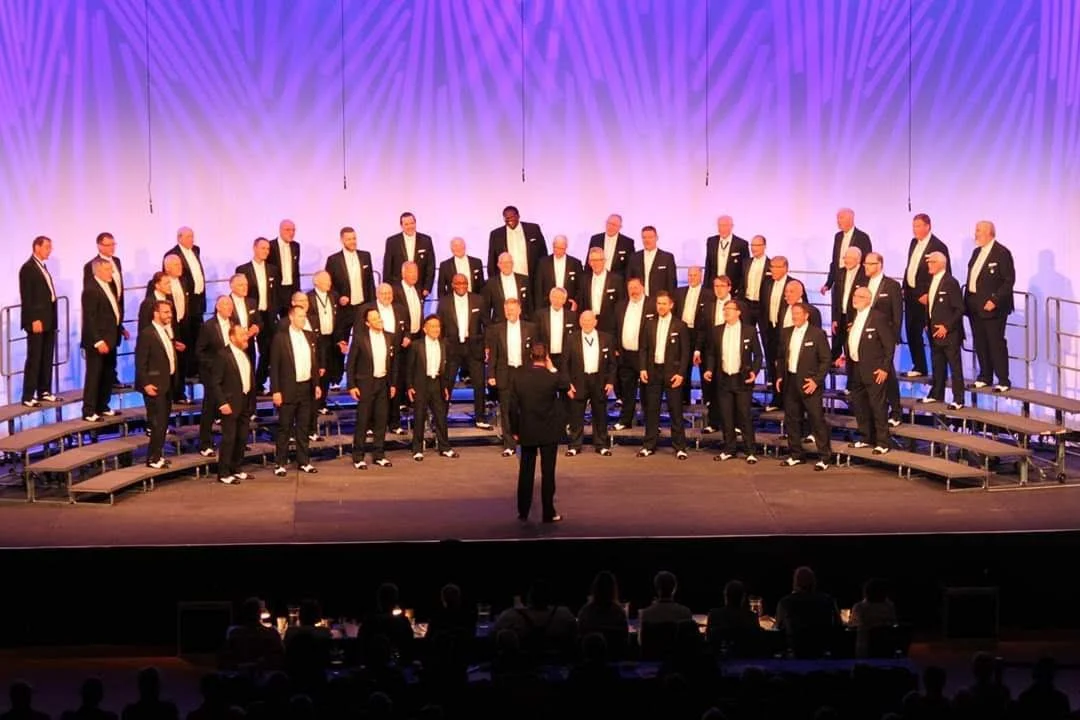 A male choir singing on stage with a conductor in front of them, all dressed in formal black suits and white shirts, with some wearing ties. The choir is arranged on tiered risers, and there is a purple and blue light pattern on the curtain backdrop.