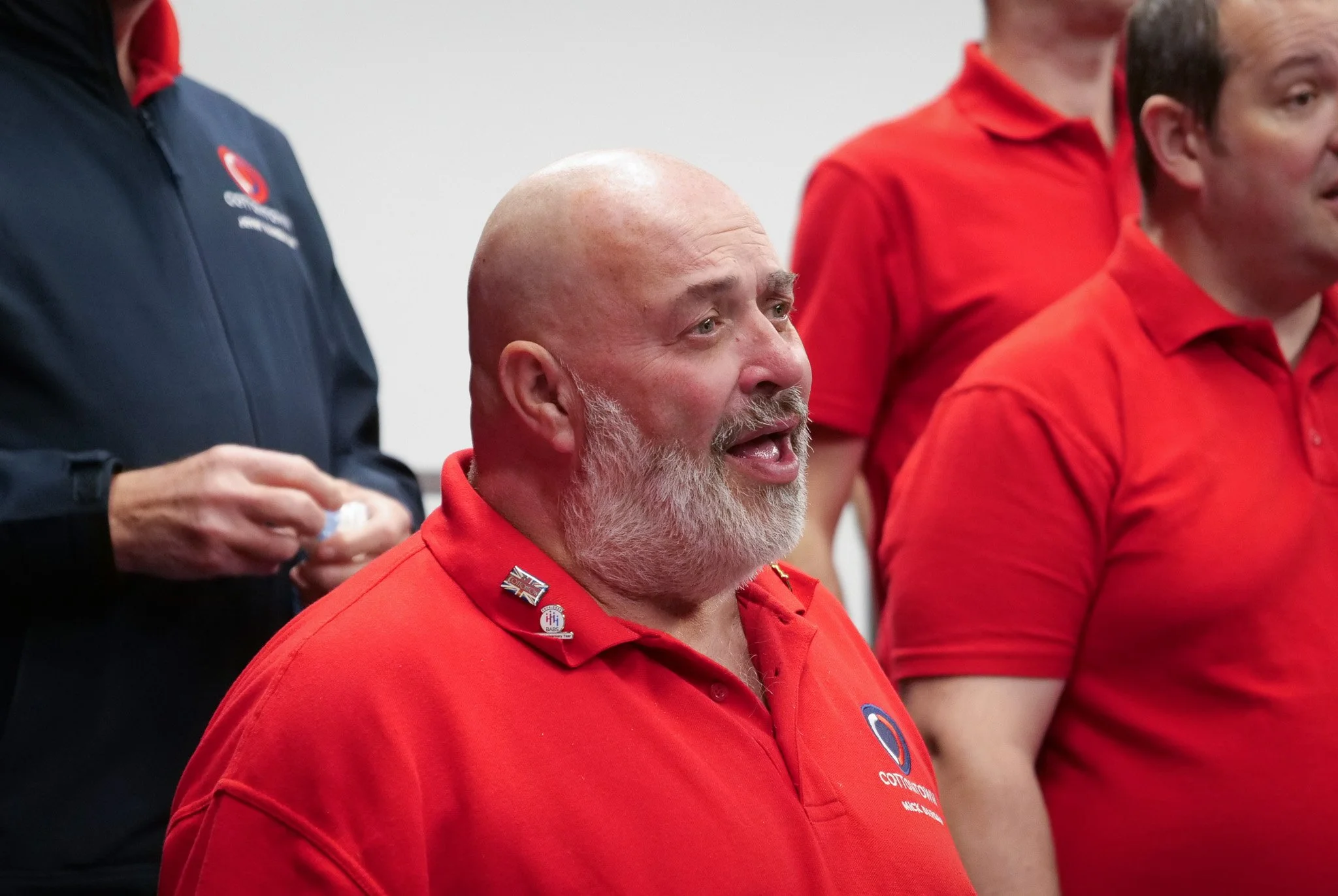 A group of men wearing red shirts, with one man in the foreground having a beard and speaking or singing, while others stand beside him. The shirt of the man in the foreground features badges and logos.