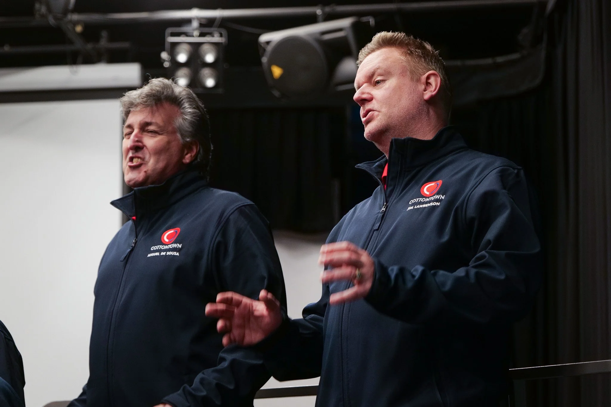 Two men wearing black jackets with the COTTONOWN logo and their names, running a presentation or speech, standing indoors with stage lights and sound equipment above.