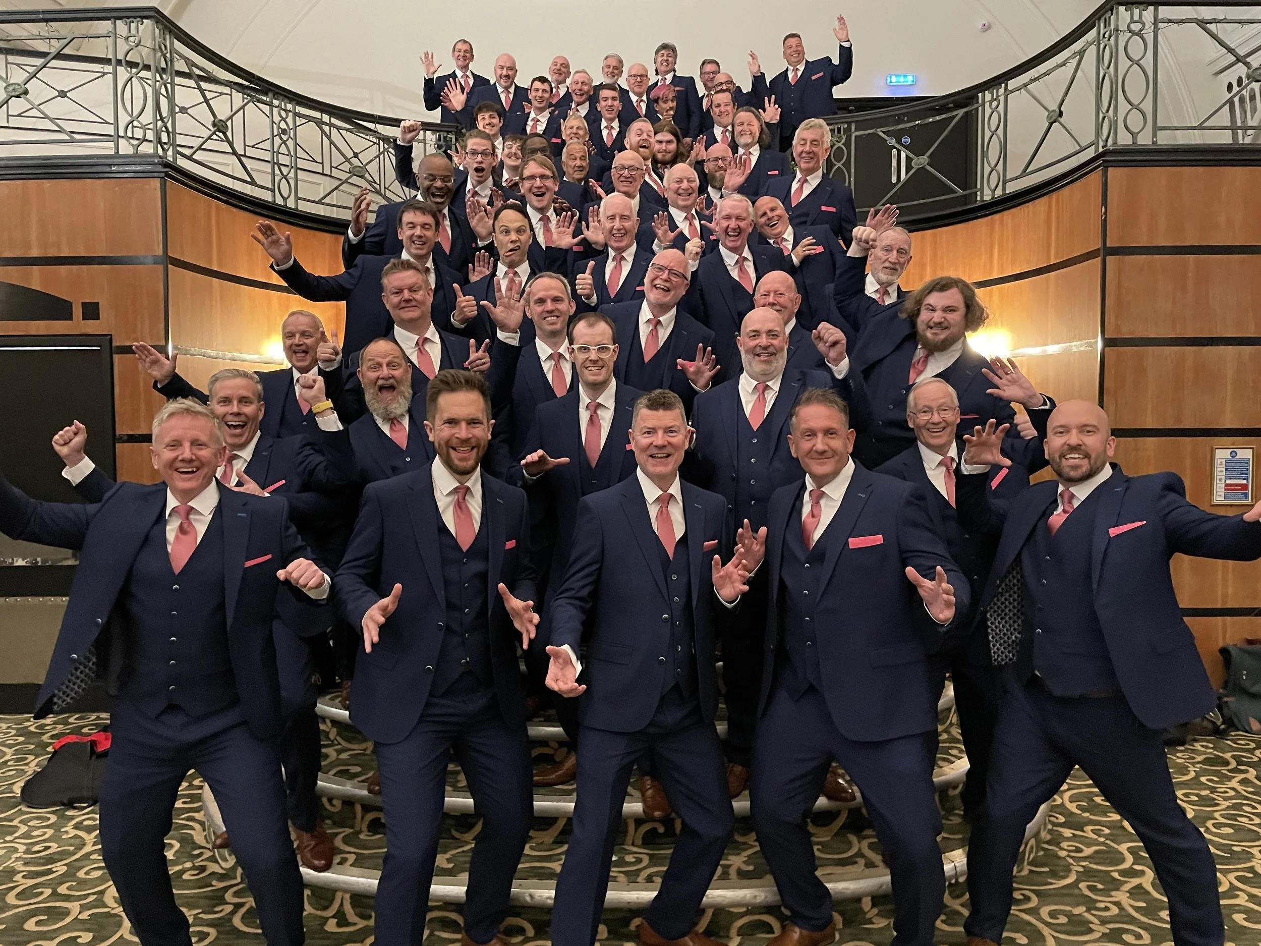 A large group of men in formal navy blue suits with pink ties and pocket squares, posing happily on a staircase in a banquet hall.