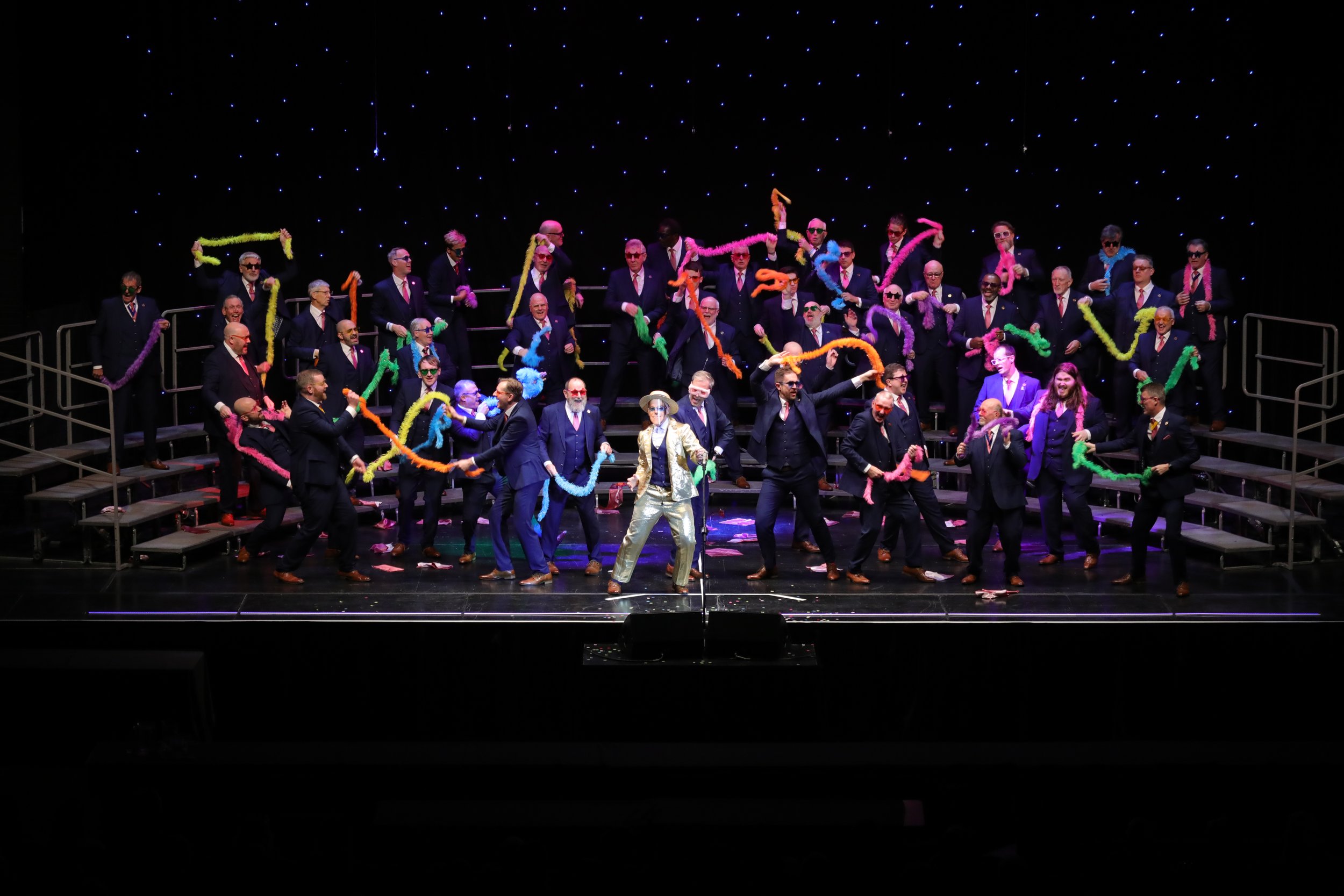 A group of performers on stage, wearing dark suits and sunglasses, dancing with colorful feather boas, with a backdrop of starry lights, and one individual in the center dressed in shiny gold pants and a patterned shirt.