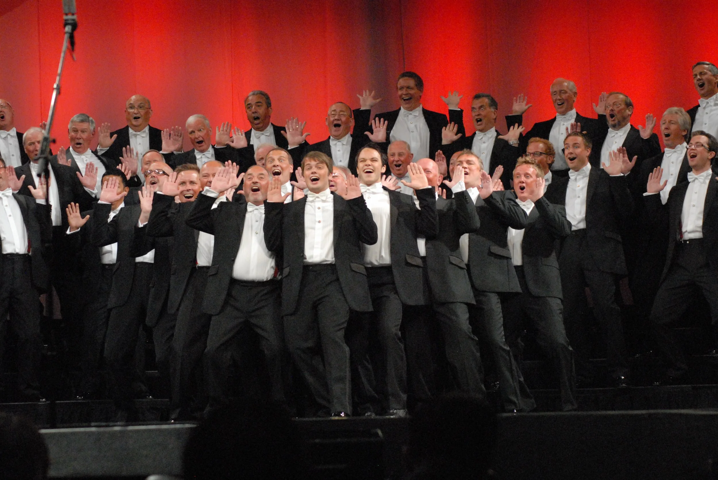 A choir of men dressed in tuxedos performing on stage with a red curtain backdrop.
