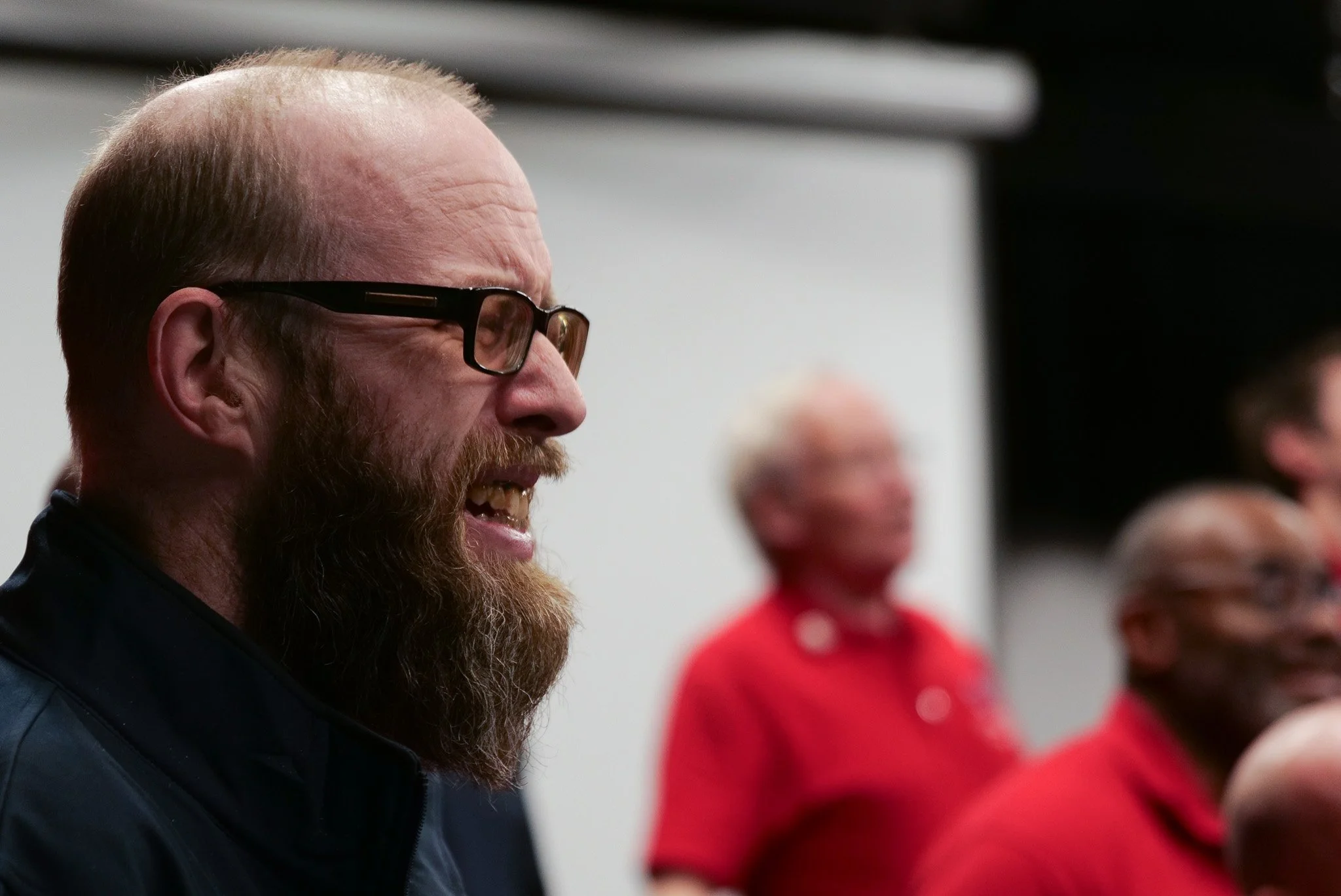A group of people, including a man with glasses and a beard, are smiling and engaging in a conversation, with two women in red shirts in the background.