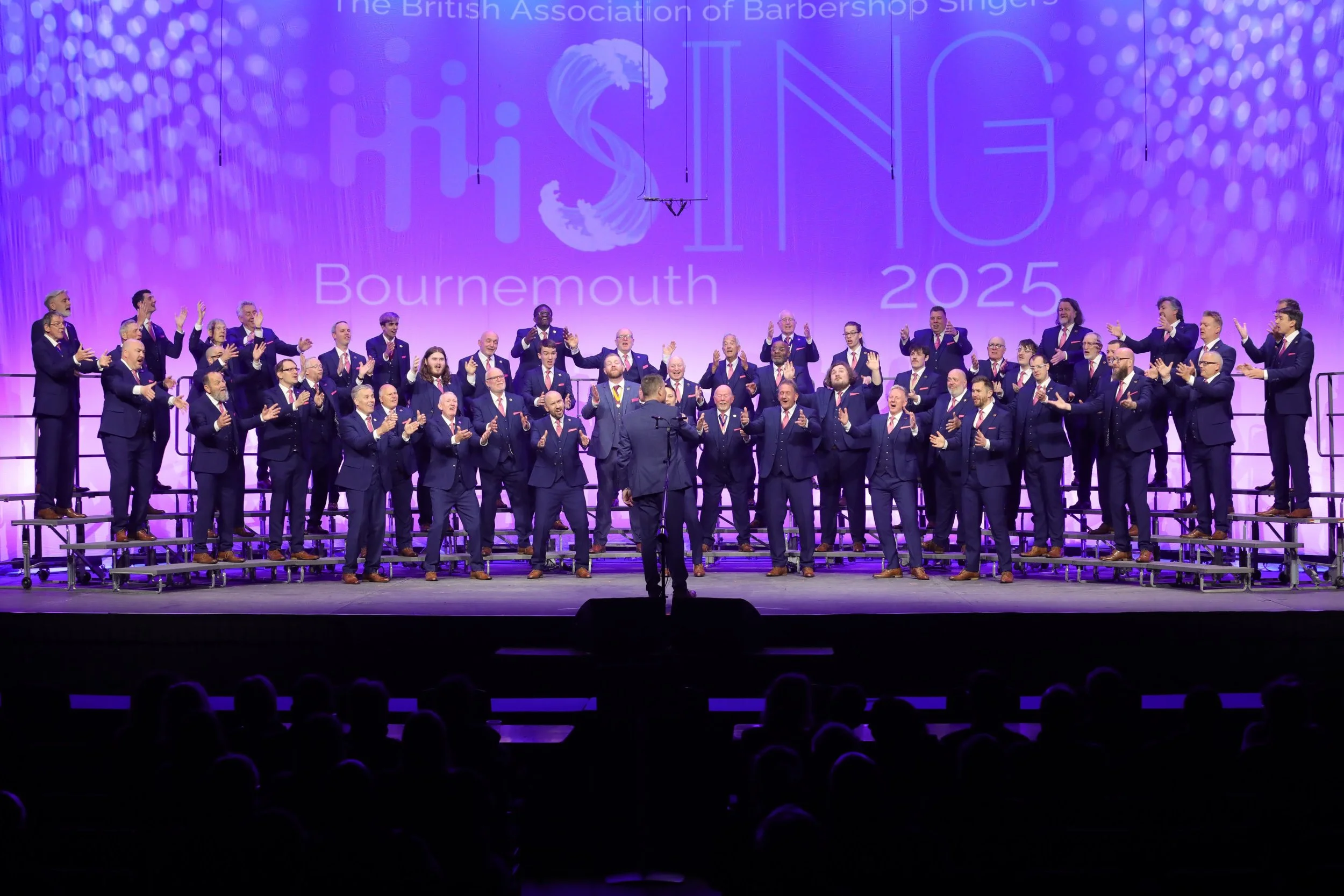 Choral group on stage performing at a conference or event. They are dressed in navy suits and standing on risers. The background displays the logo for the British Association of Barbershop Singers and the text 'HARMONY 2025 Bournemouth' with purple l