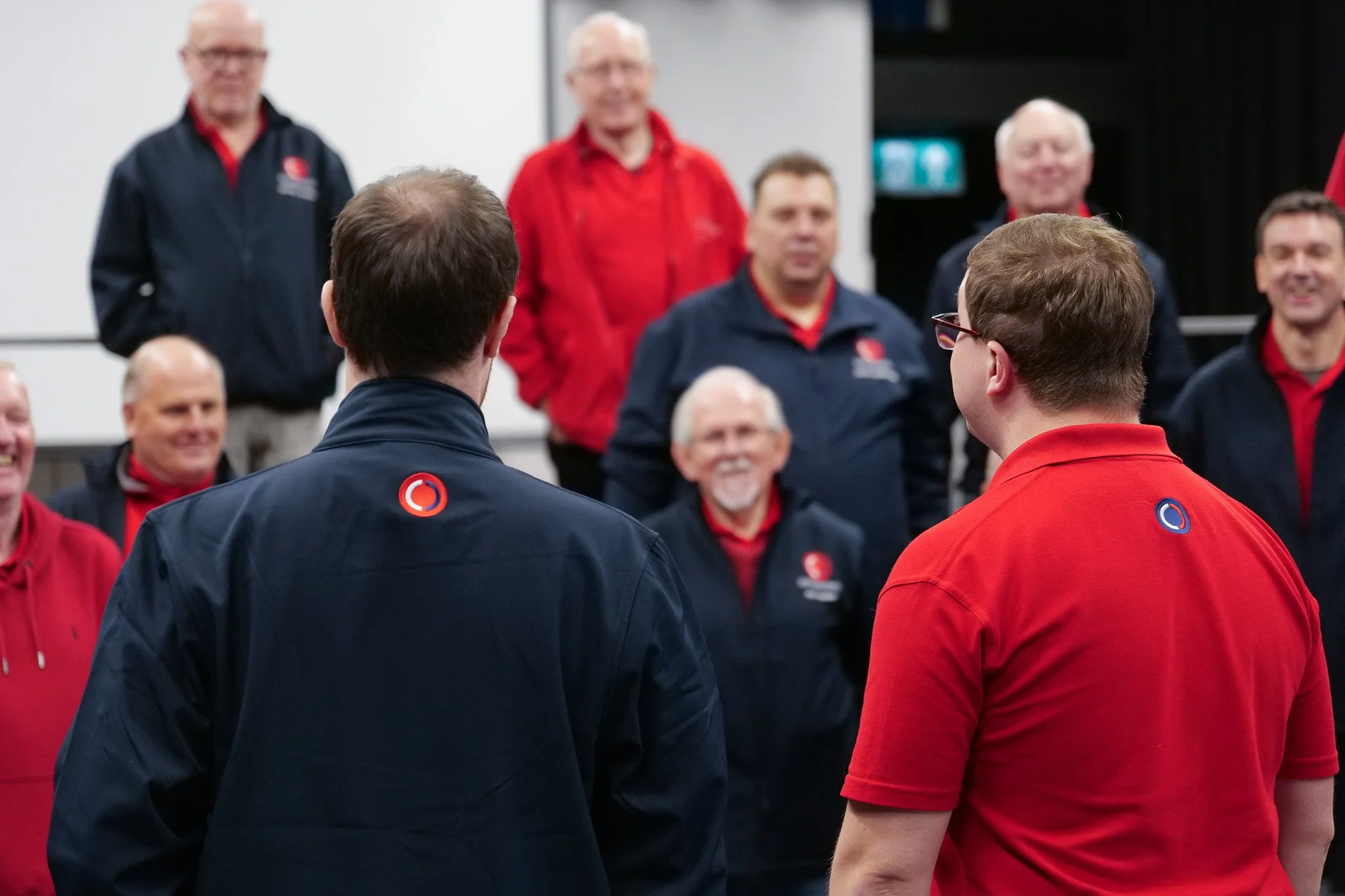 Two men with short brown hair and glasses wearing dark and red shirts, standing with their backs to the camera facing a group of smiling older men in matching dark and red jackets, in an indoor setting.