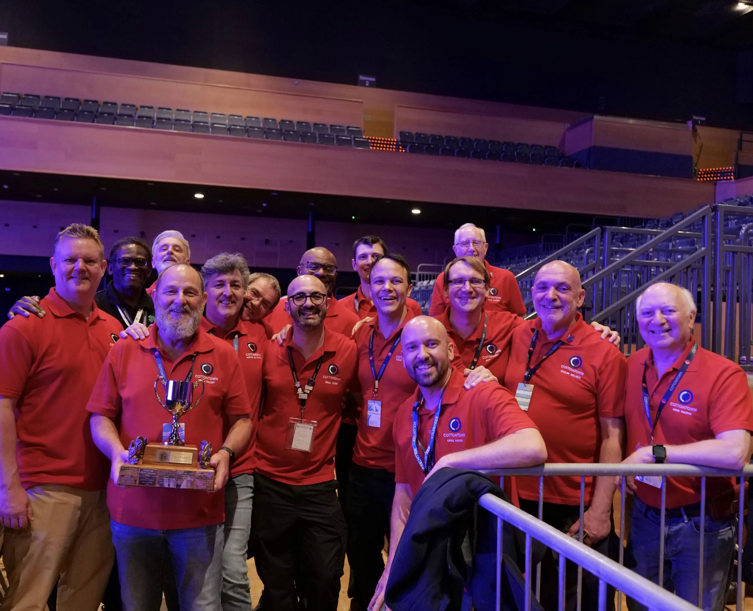 Group of men in red shirts at a barbershop conventio , some holding a trophy, inside an auditorium.