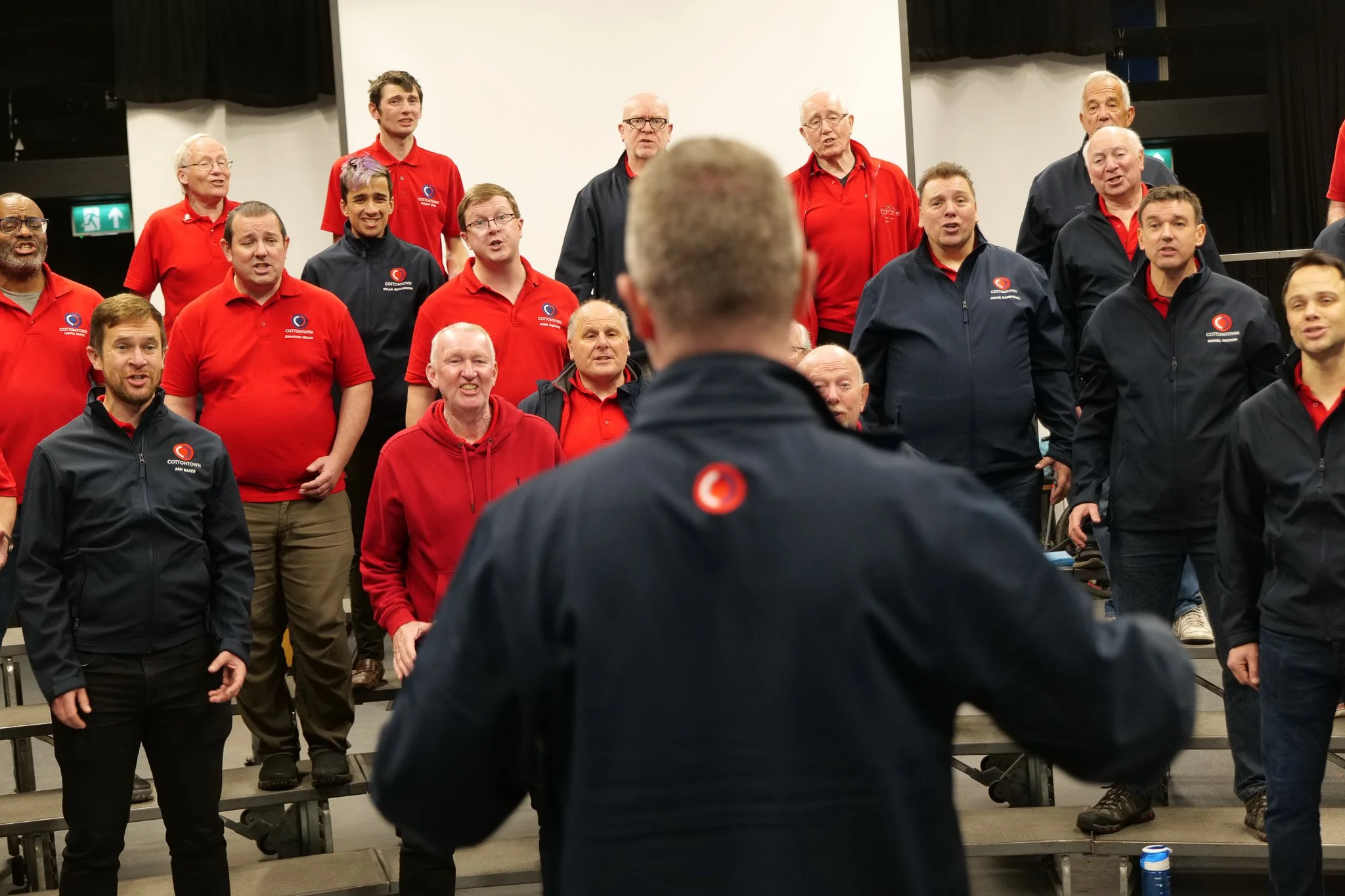 A group of men and women singing or performing in a choir, viewed from behind a conductor.
