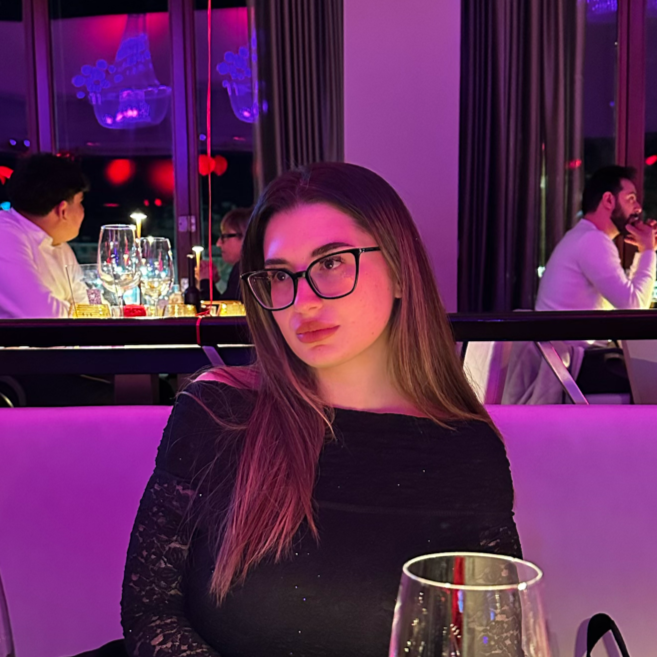 Young woman with long brown hair and glasses sitting at a restaurant table with purple lighting, surrounded by other diners and glassware.