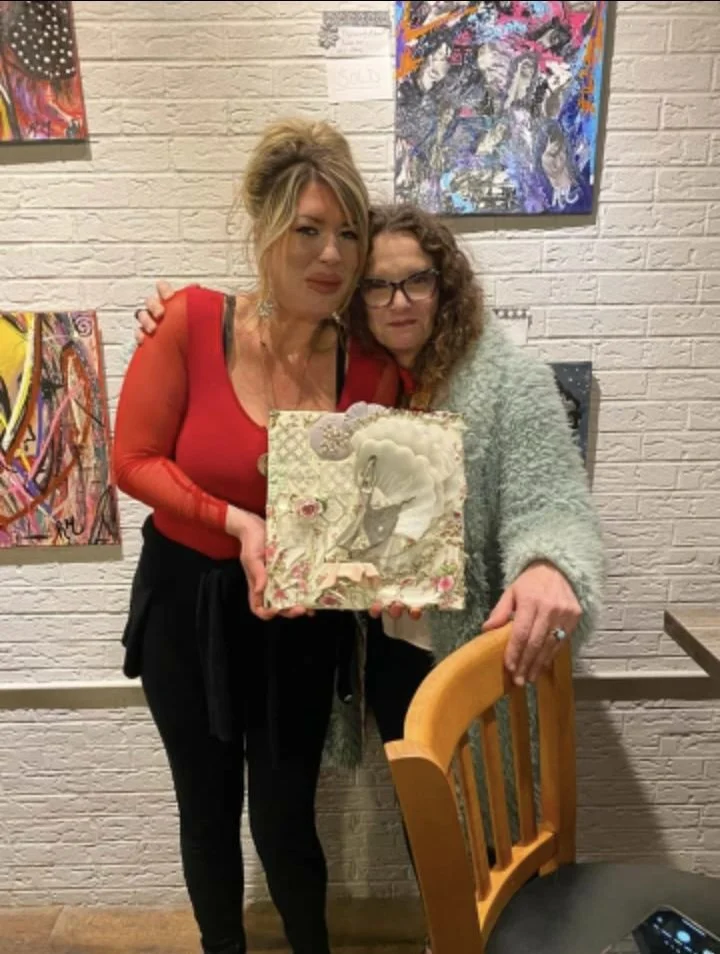 Two women stand close together, holding a decorative art piece with a swan illustration. They are inside an art gallery, with colorful abstract paintings on the brick wall behind them.