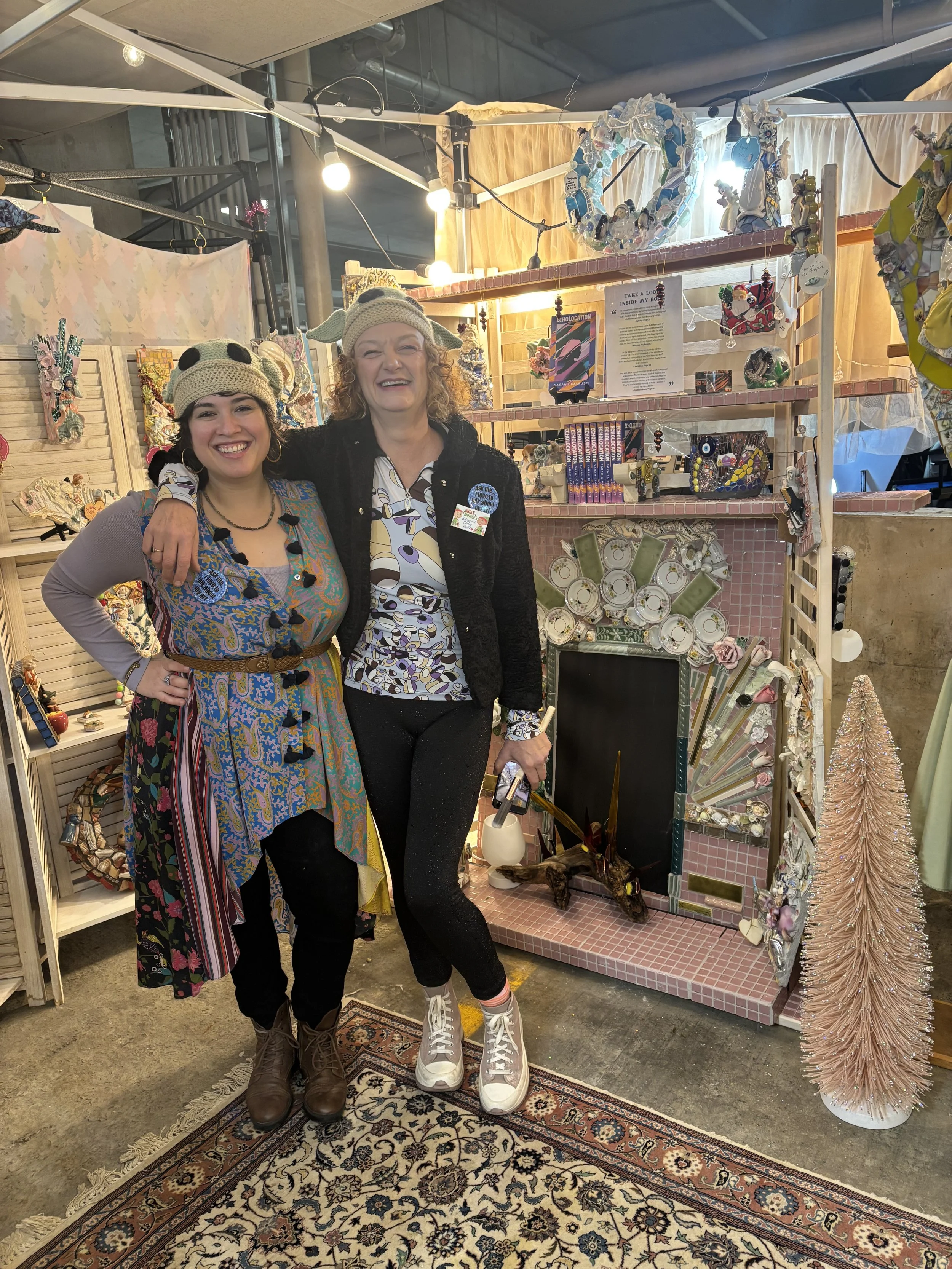 Two women standing together, smiling, at a craft fair booth decorated with various handmade items. One woman is wearing a panda hat, and the other is wearing a hat with a pom-pom and a button. The booth features shelves with ceramics, artwork, and ornaments, and a pink tiled fireplace with a unicorn sculpture.