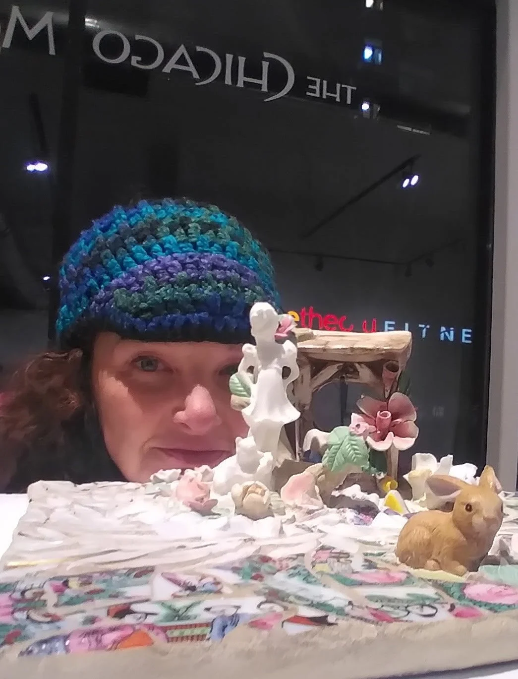 Woman with a colorful knitted hat behind a display of ceramic animal figurines, including a rabbit, and flowers, with a dark storefront background.