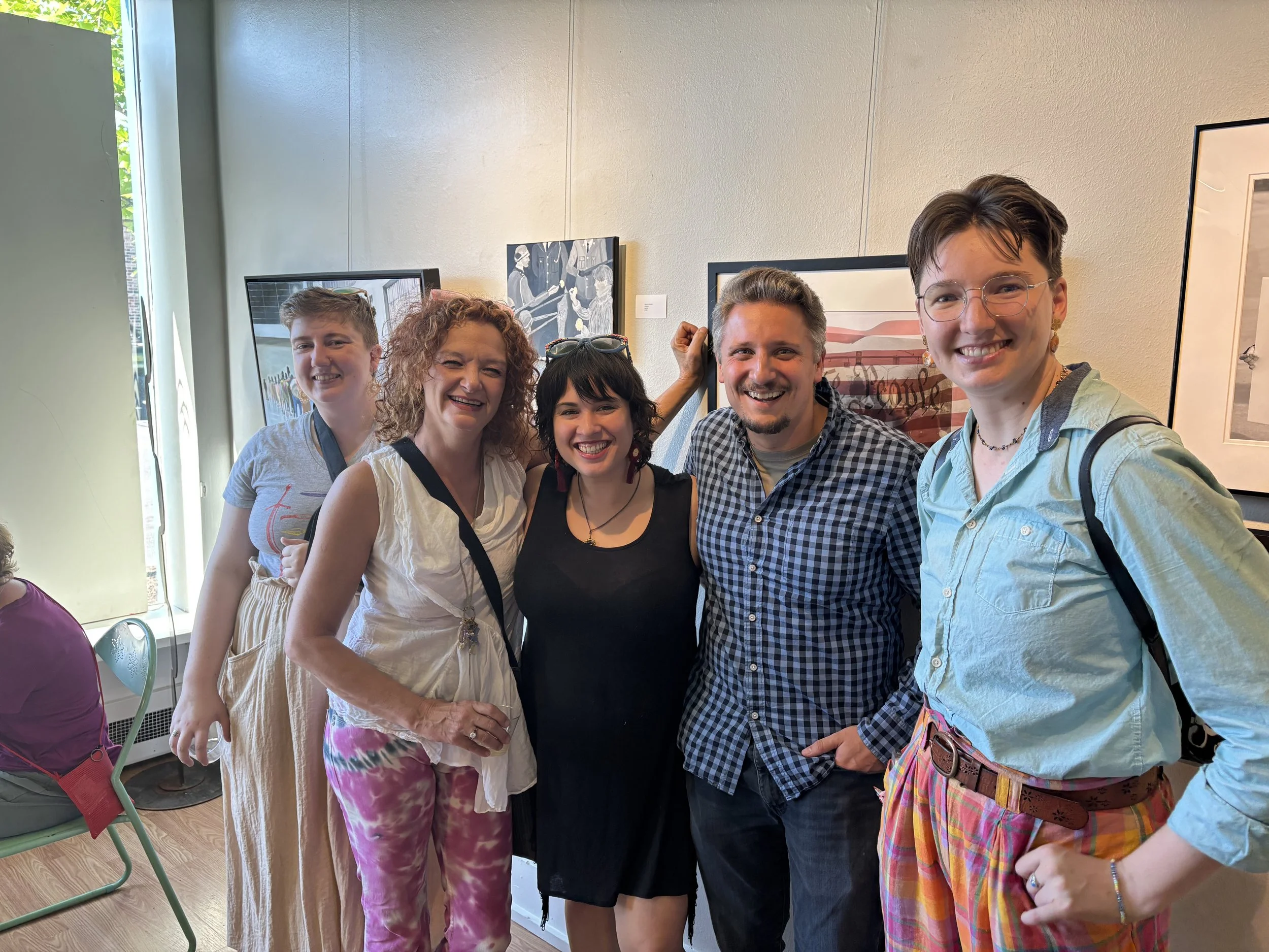 Five people smiling and standing together in an art gallery with framed artwork on the wall behind them.