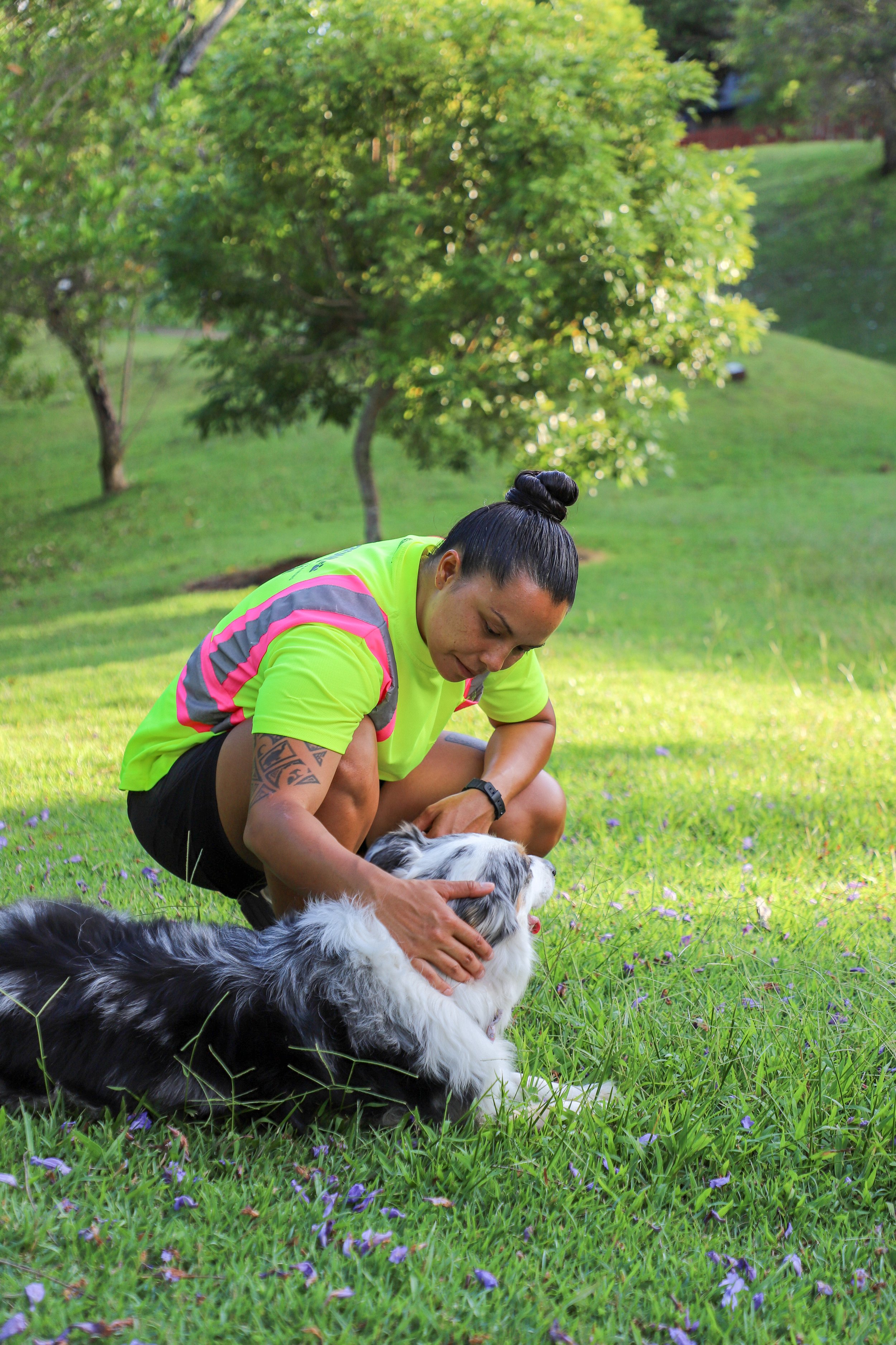 A woman in a bright yellow athletic shirt and black shorts kneeling on grass, petting a black and white Australian Shepherd dog, with green trees and a grassy yard in the background.