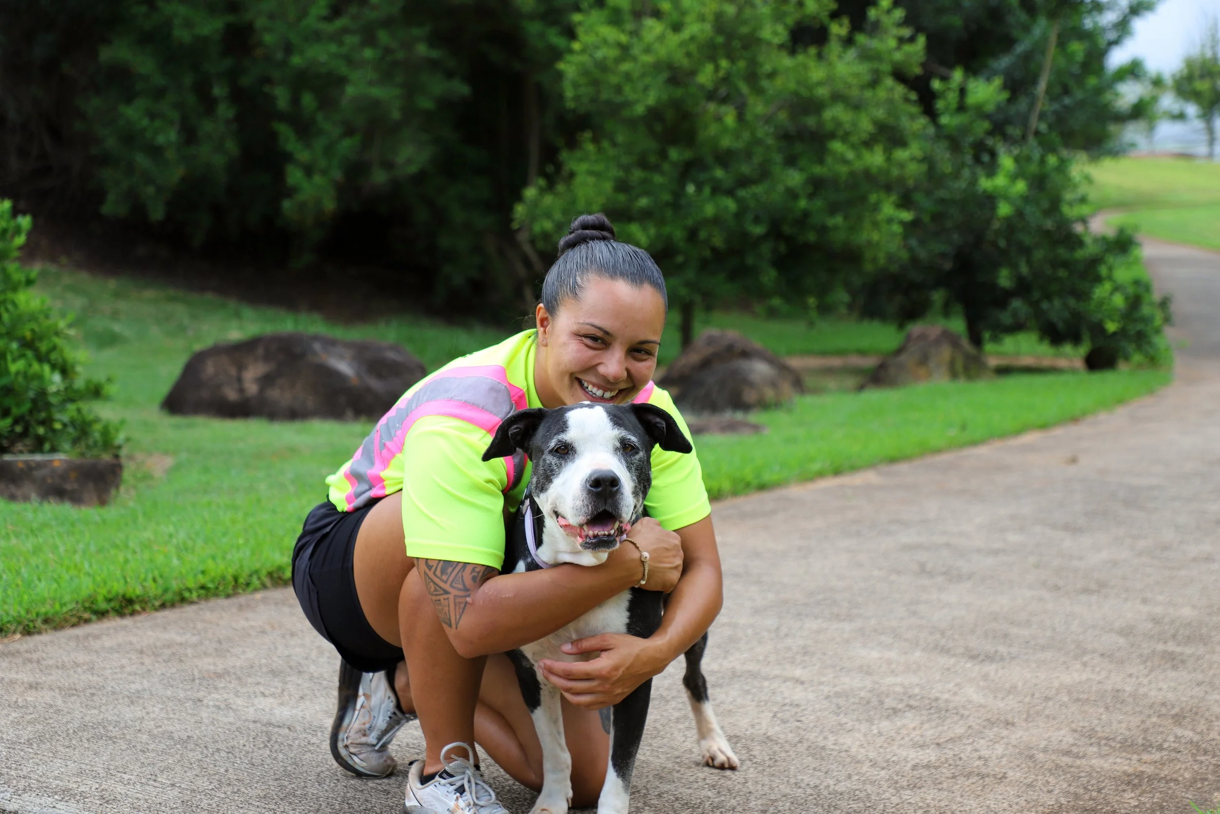 A woman hugging a black and white dog outdoors in a park with green trees and rocks in the background.