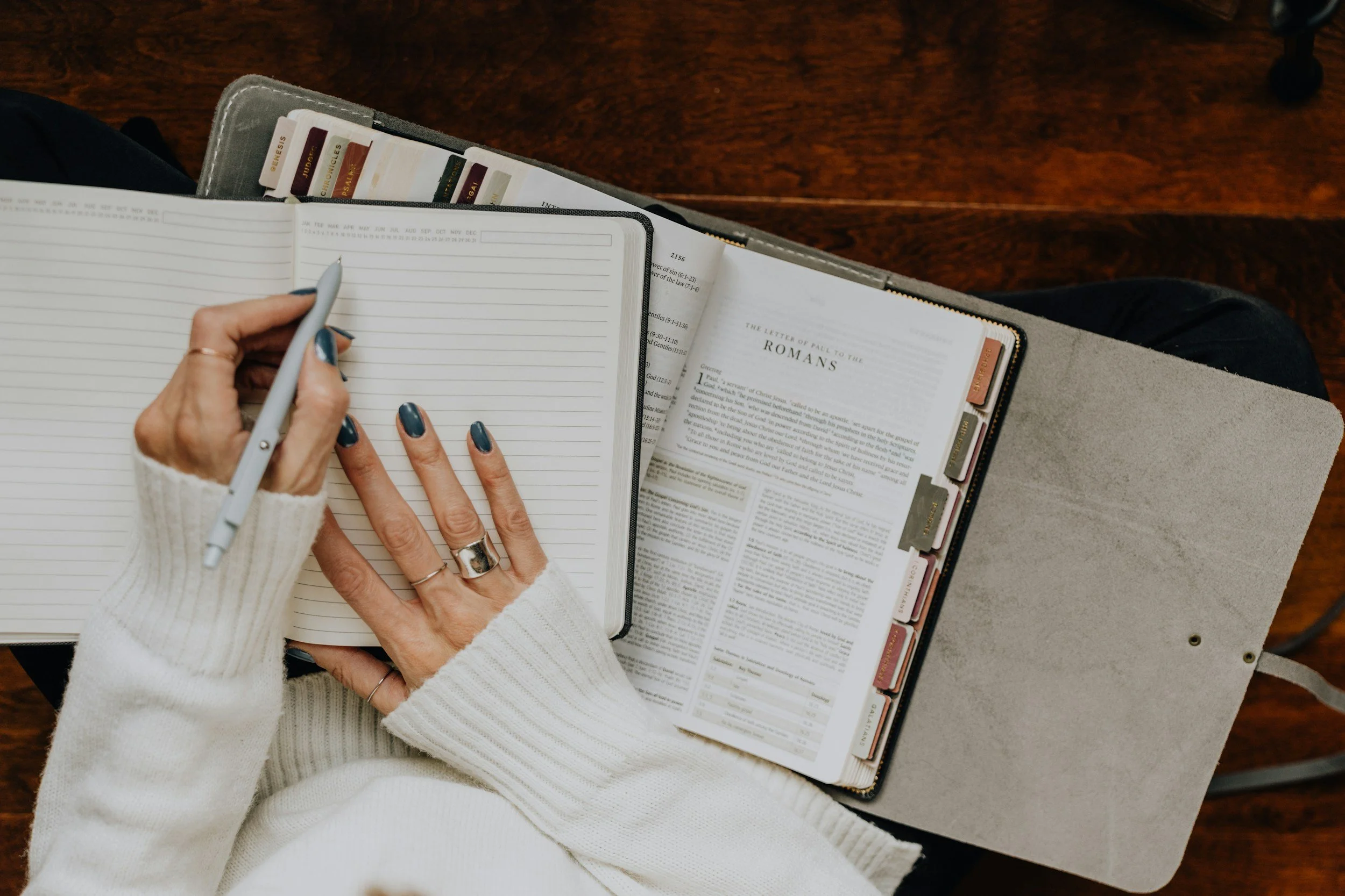 Person with painted nails and rings on hand writing in a lined notebook, with open Bible showing Romans.