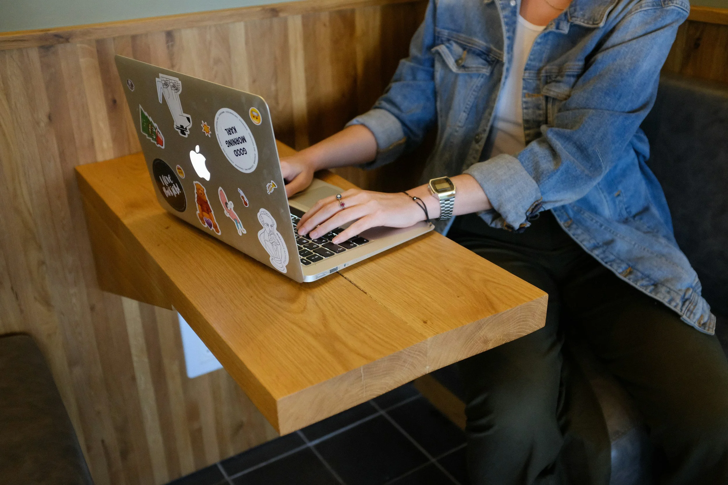 Person sitting at a wooden table using a laptop with stickers on the cover, wearing a denim jacket and a watch.