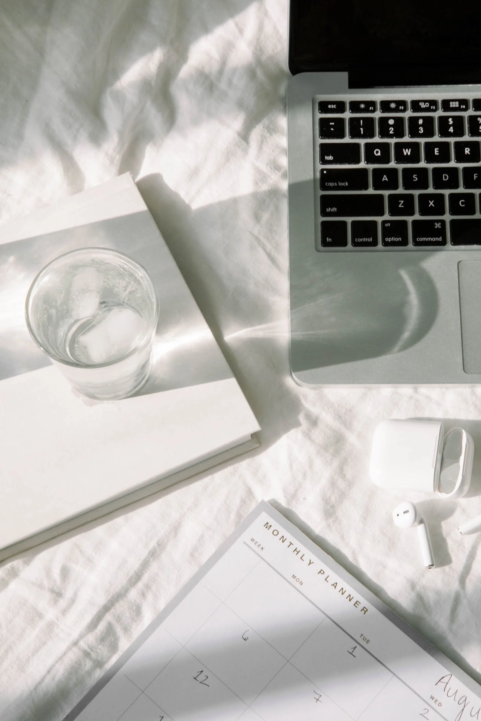 Laptop computer, glass of water with ice on a white notebook, wireless earbuds and case, monthly planner, all on a white fabric surface.