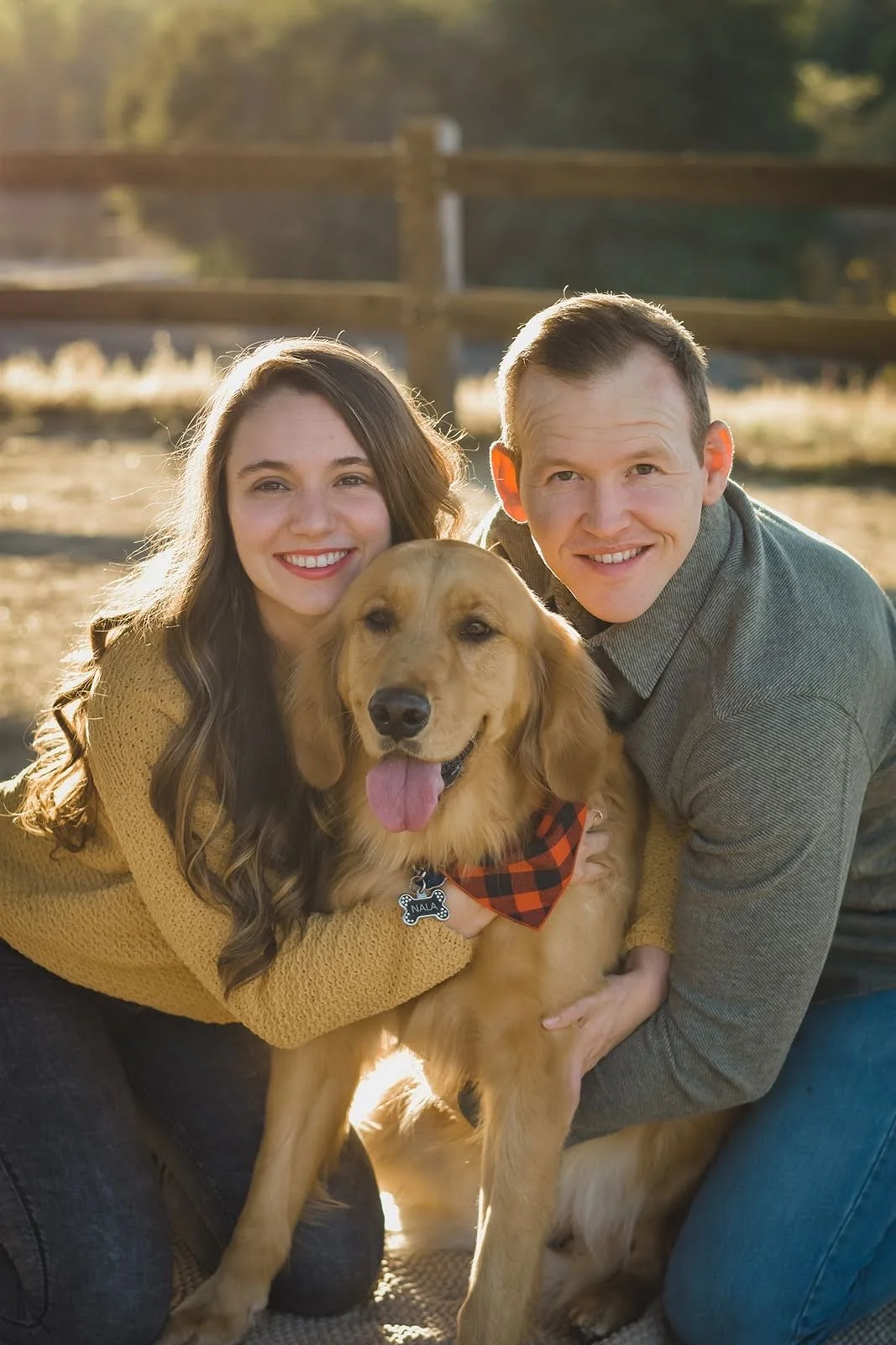 A smiling young woman and a smiling young man are hugging a happy golden retriever dog outdoors during golden hour, with a wooden fence in the background.