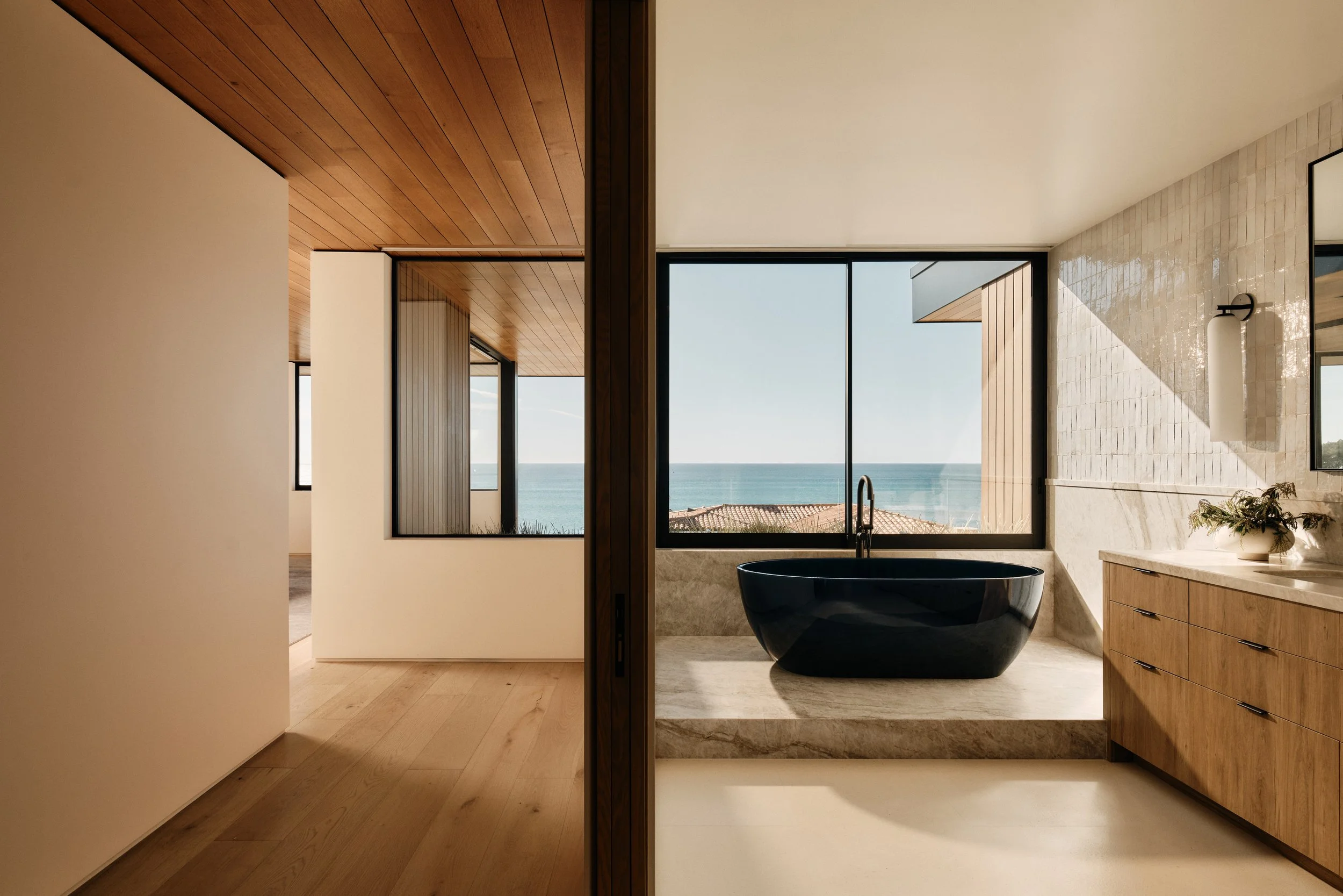Modern bathroom with a bathtub near a large window overlooking the ocean, wooden cabinets, and handmade tiled walls.