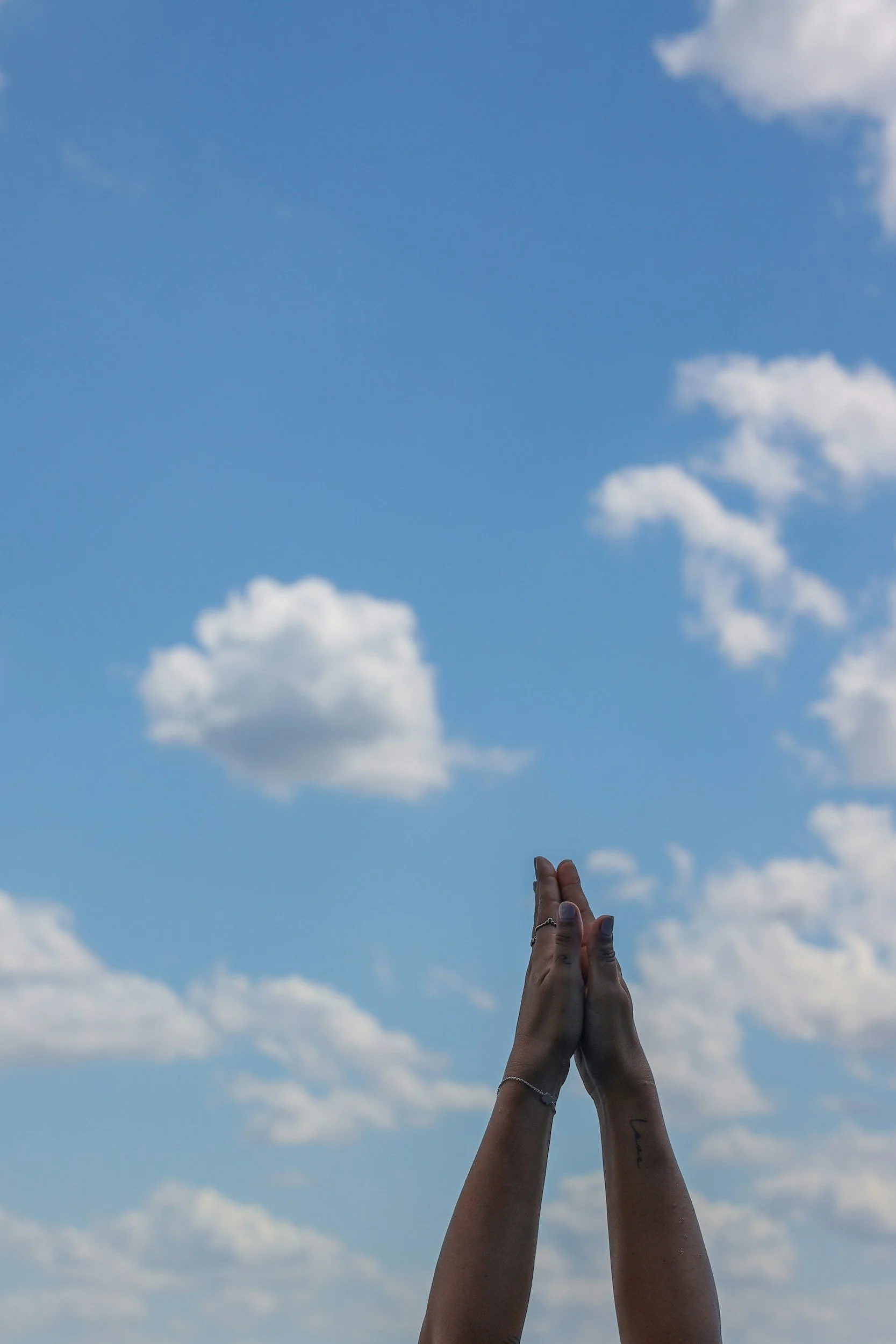 A pair of hands pressed together in a prayer or greeting gesture against a bright blue sky with scattered white clouds.