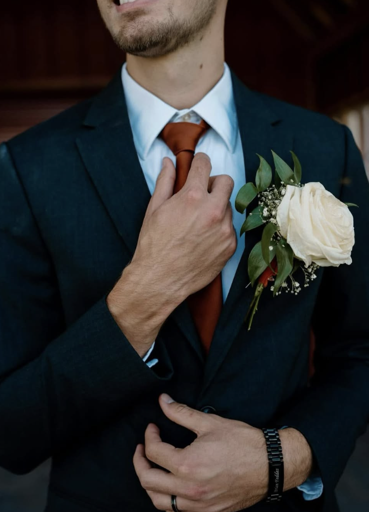 A man dressed in a dark suit with a white shirt and a reddish-brown tie, with a white rose boutonniere with Italian ruscus leaves on his lapel. He's adjusting his tie with one hand and wearing a bracelet and a ring.