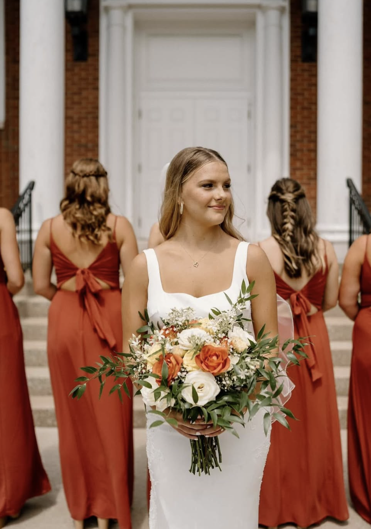 A bride in a white wedding dress holding a bouquet of flowers, standing in front of a group of bridesmaids in rust orange dresses on the steps of a building.