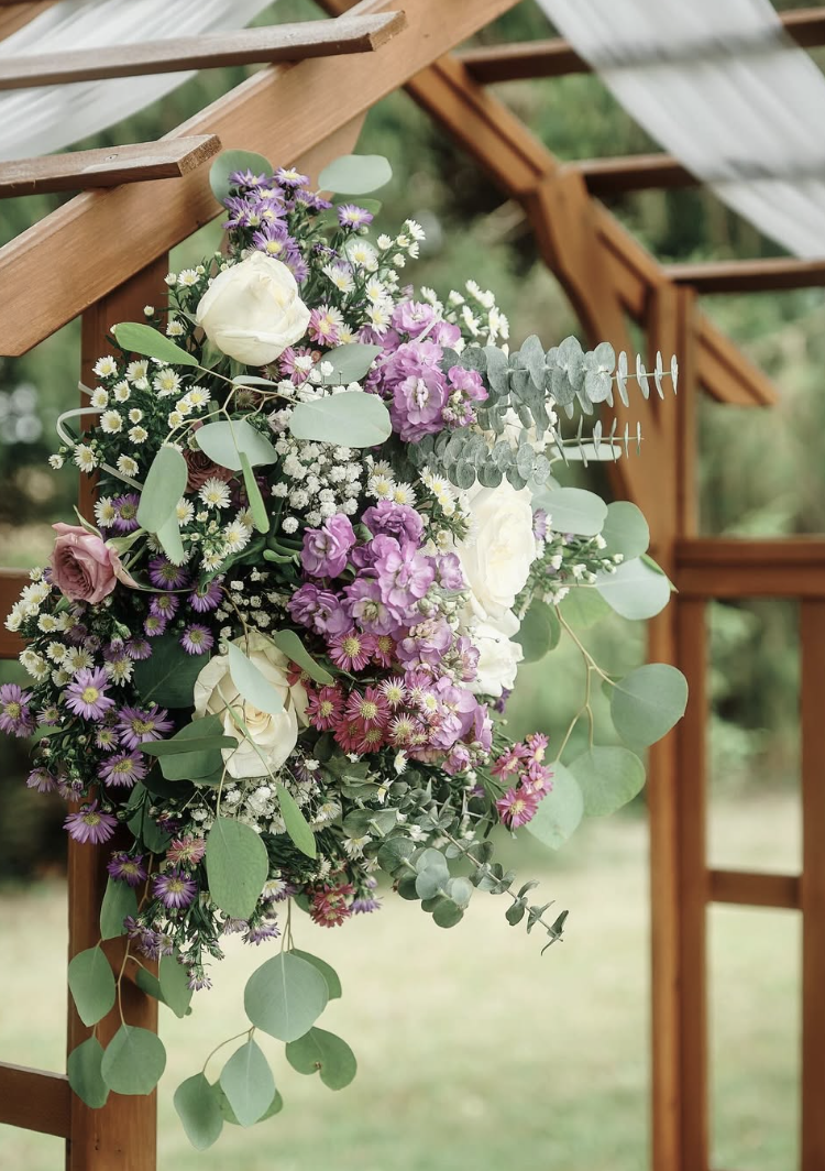 Floral arrangement with white roses, purple and pink flowers, baby's breath, and eucalyptus leaves attached to wooden outdoor structure.