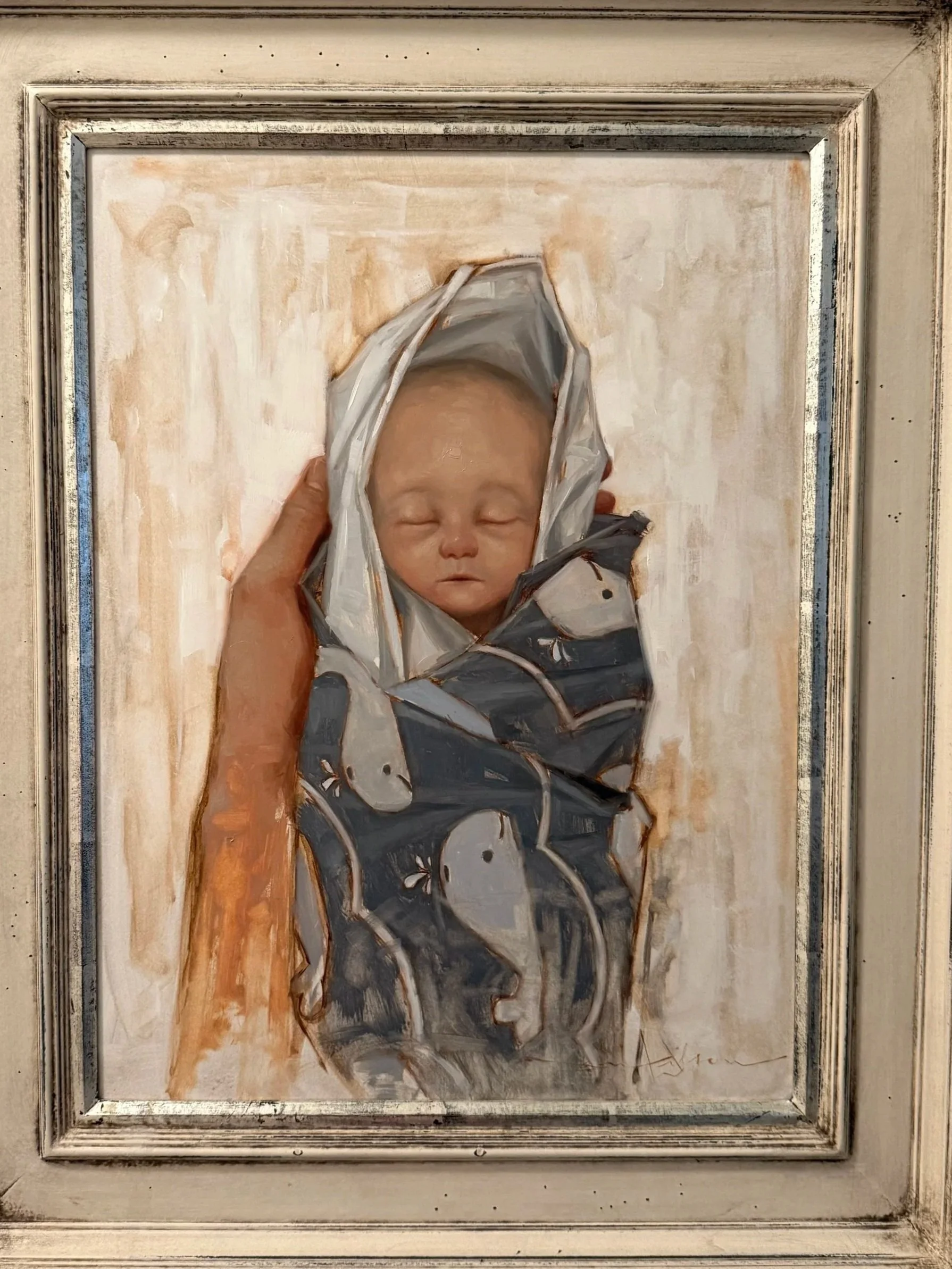 Painting of a sleeping baby wrapped in a blanket, held by a hand, set in a decorative frame.