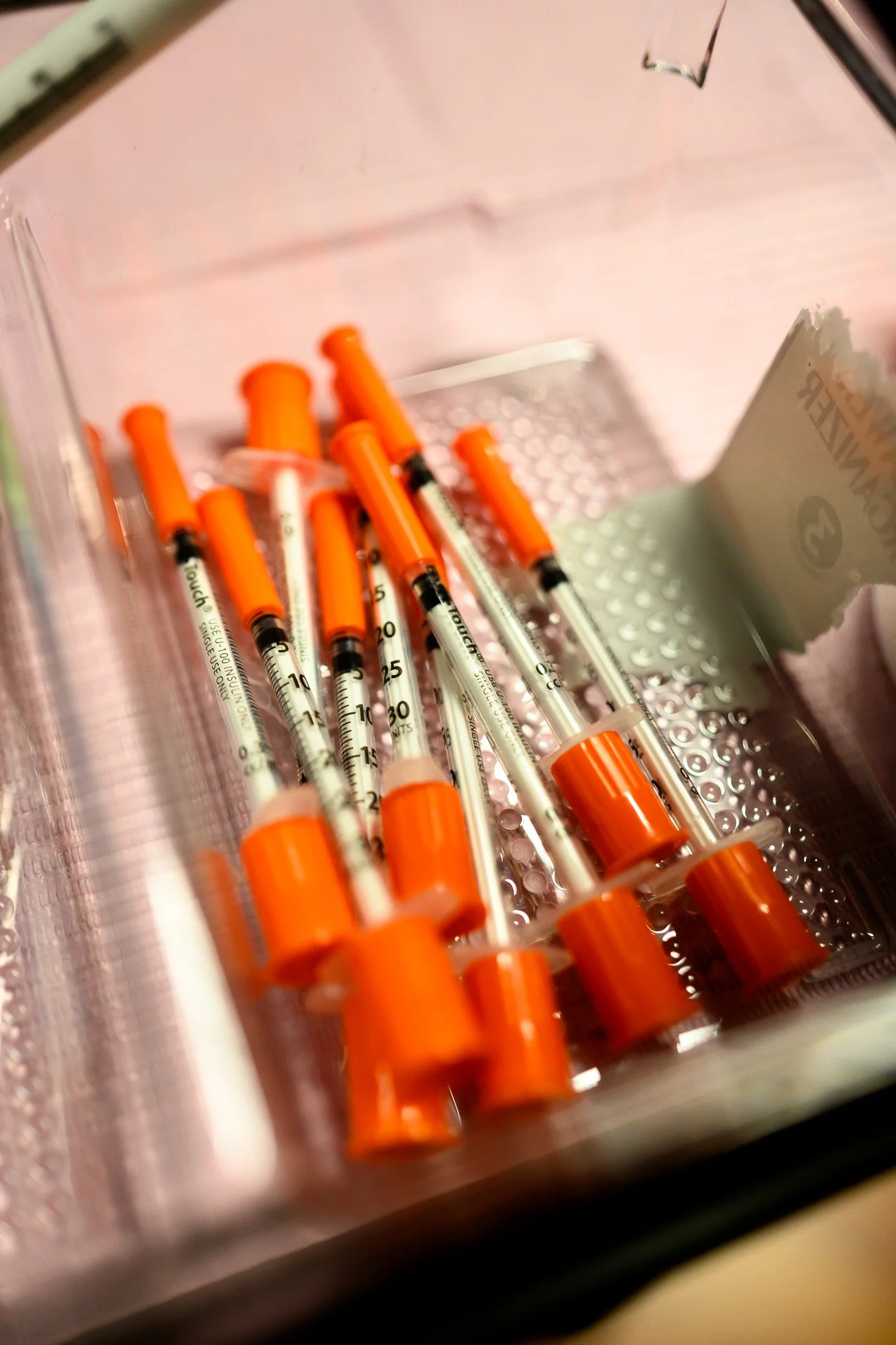 Multiple syringes with orange caps inside a plastic container, with a roll of medical tape or gauze in the background.