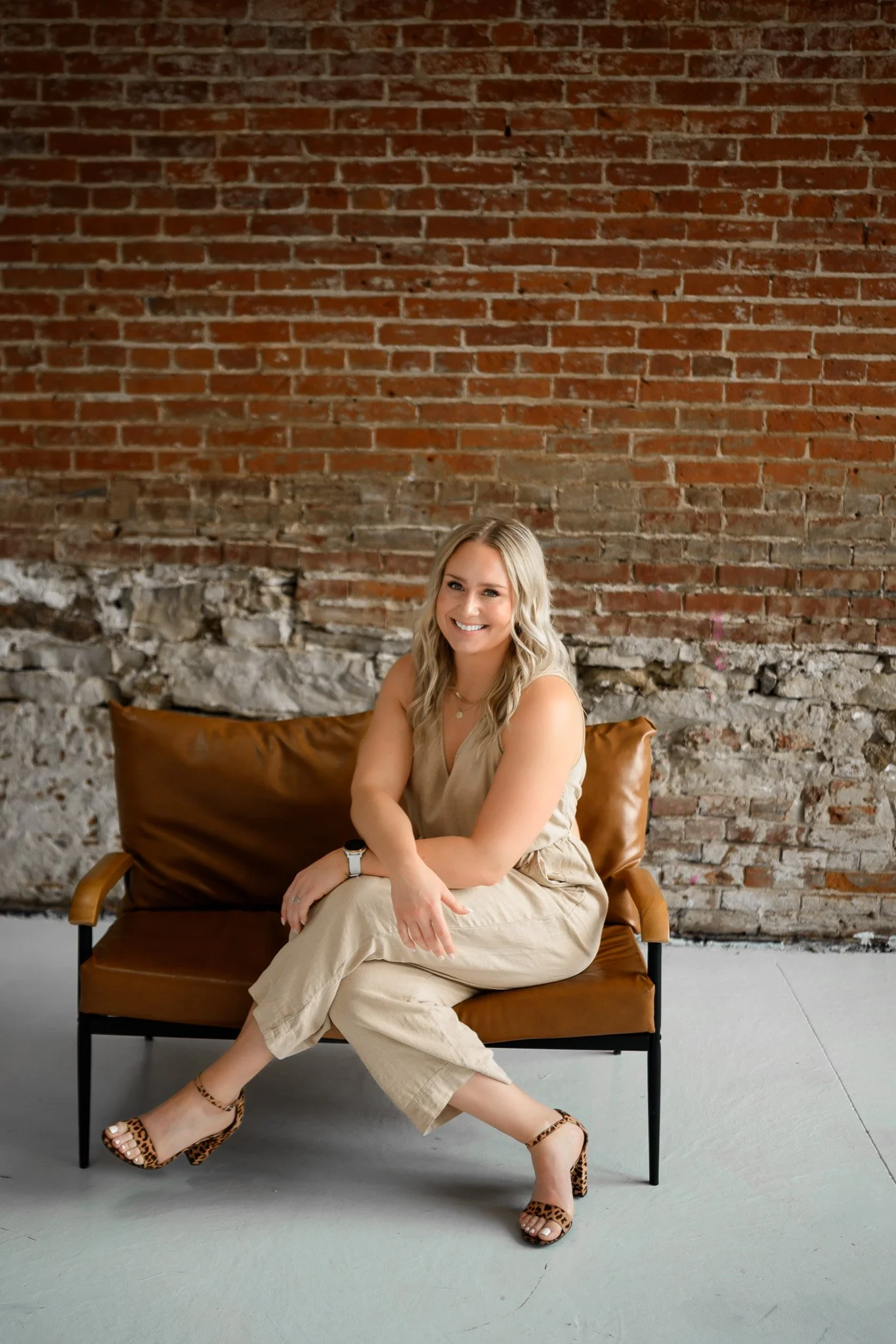 A woman with blonde hair sitting on a brown leather sofa against a brick wall, smiling at the camera, wearing a beige outfit and leopard print heels.