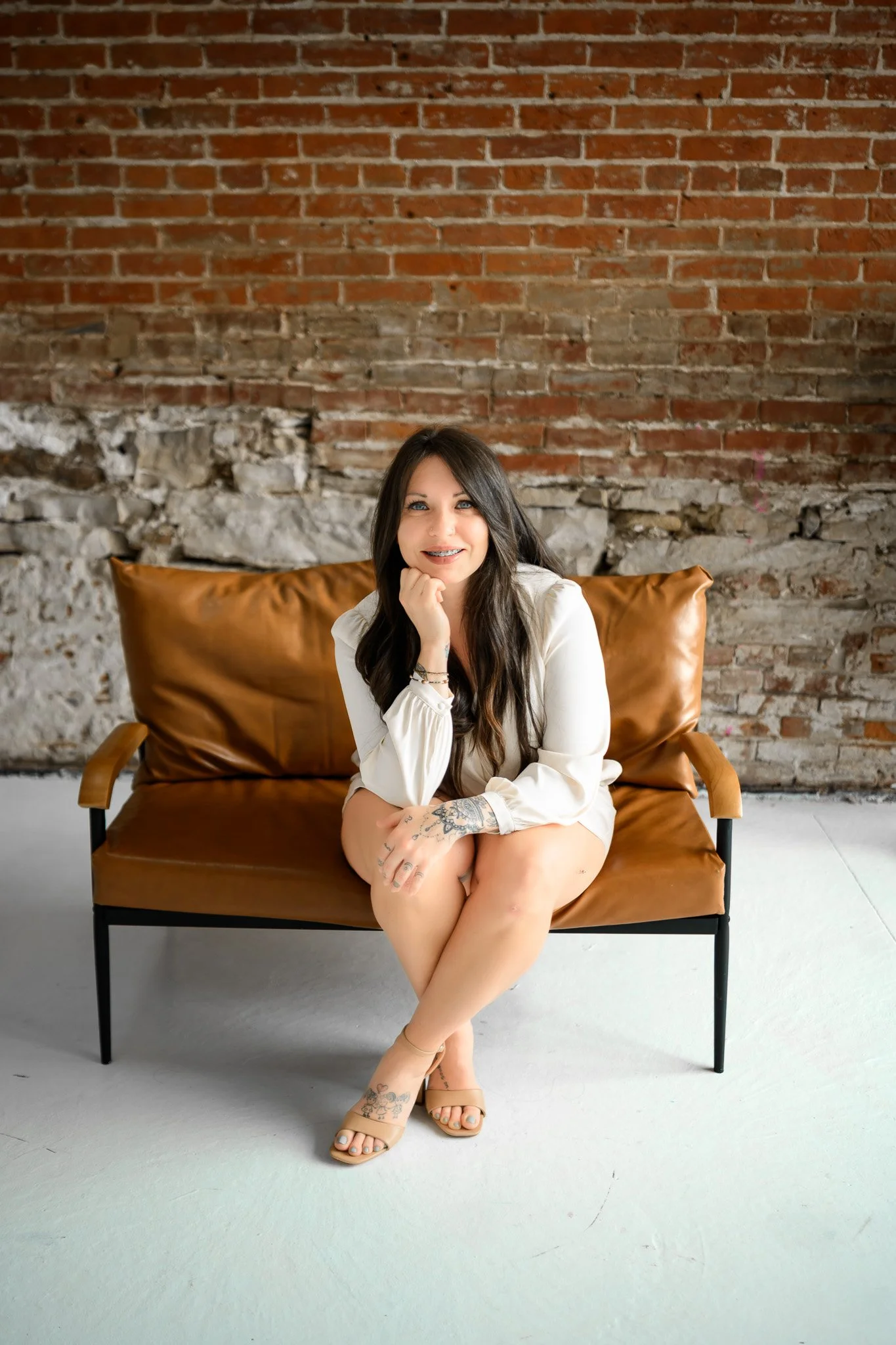 A woman with dark hair and tattoos on her hand and foot, sitting on a brown leather sofa in front of a brick wall, smiling at the camera.
