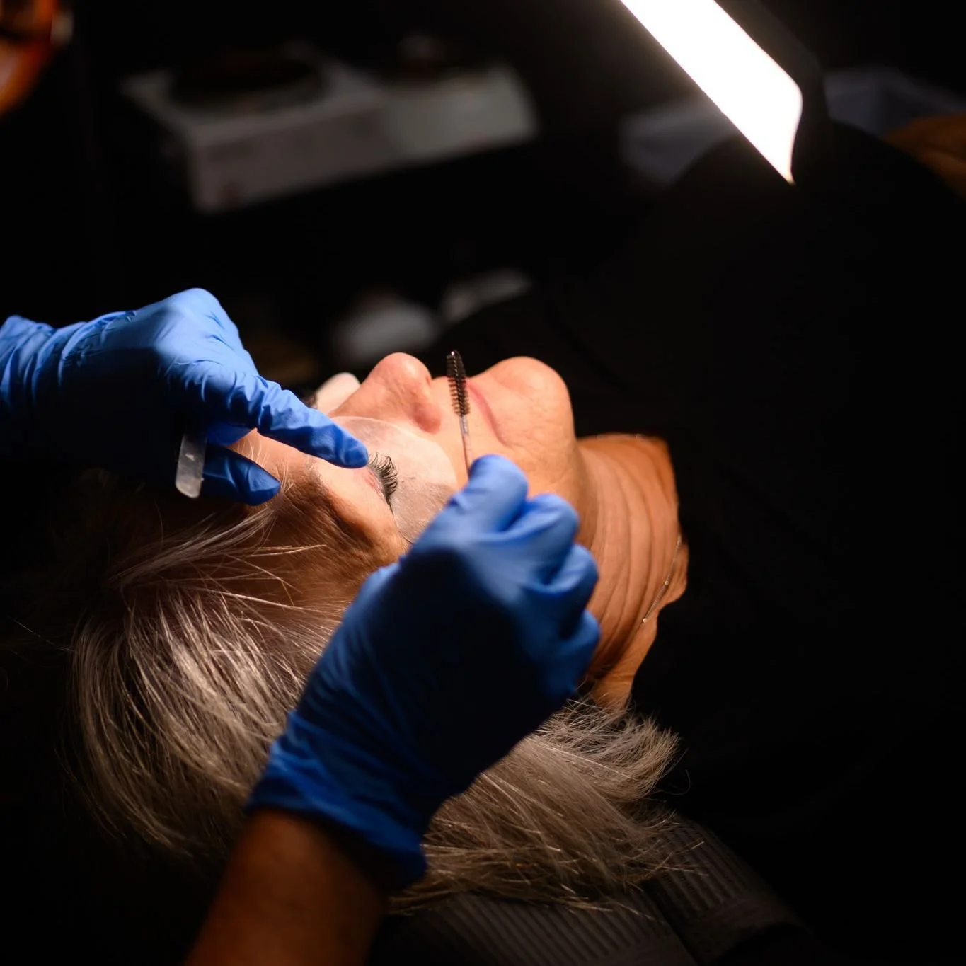 A person receiving an eyebrow grooming treatment with a microblading tool, while lying down with closed eyes. A technician in blue gloves is working on the person's eyebrows under a bright light.