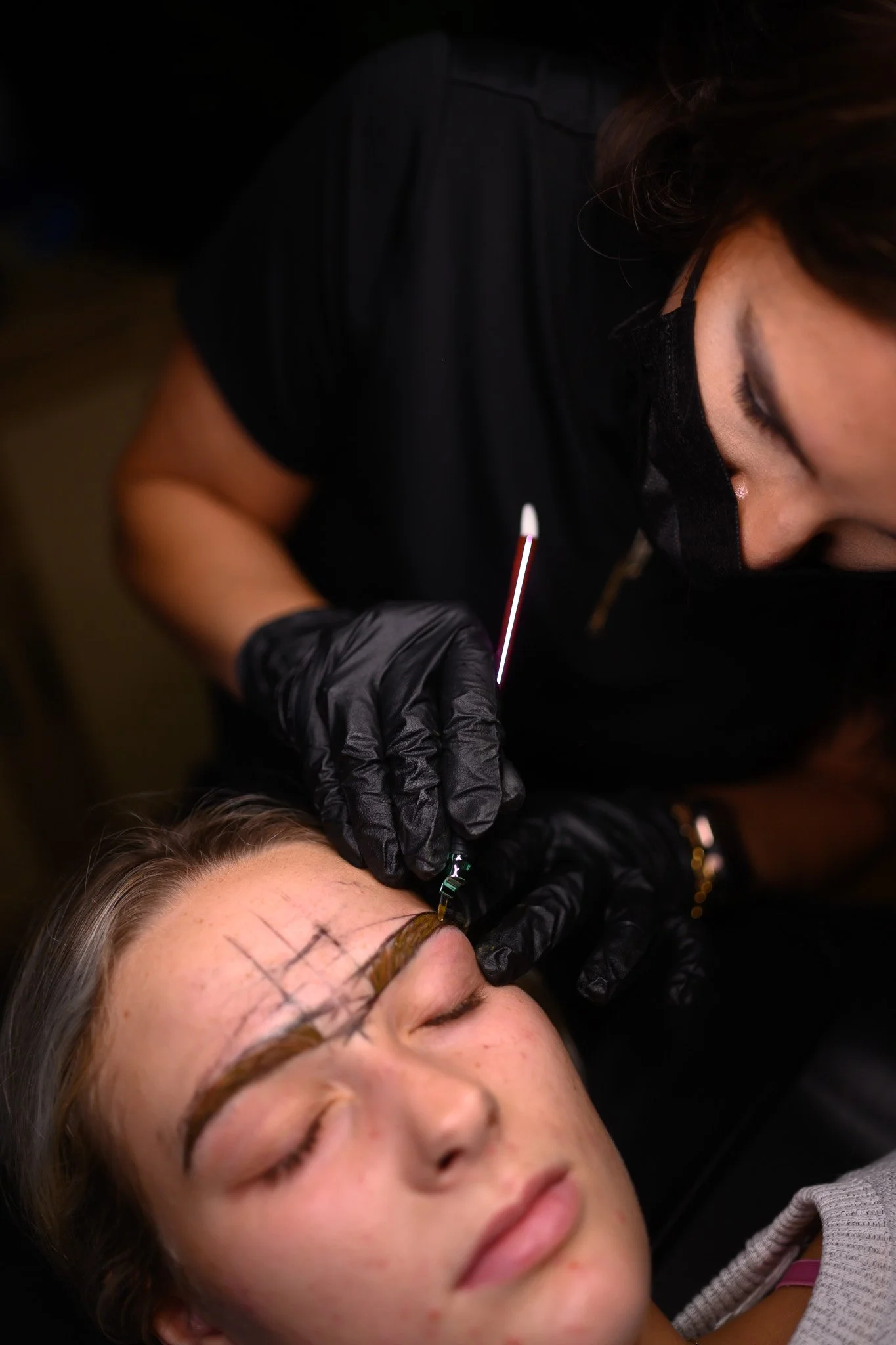A woman is getting her eyebrows tattooed or microbladed by a professional wearing black gloves and a black mask.