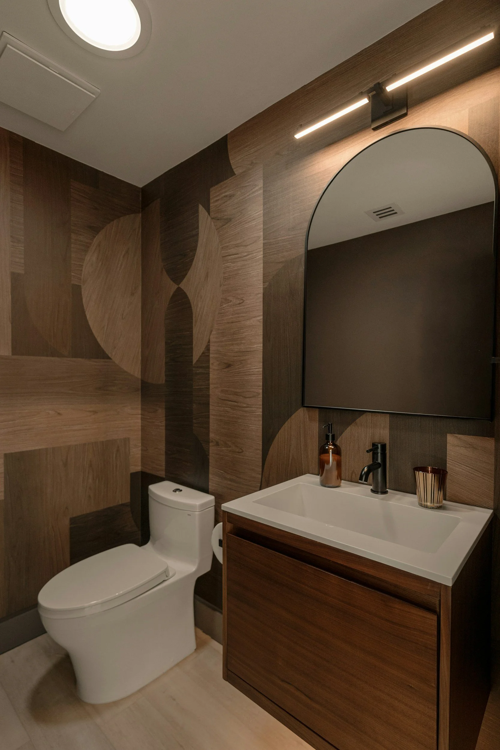 Modern bathroom with wood-paneled walls, a toilet, a wooden vanity with a white sink, a black faucet, a soap dispenser, a cup, and a large arched mirror. Overhead lighting is present.