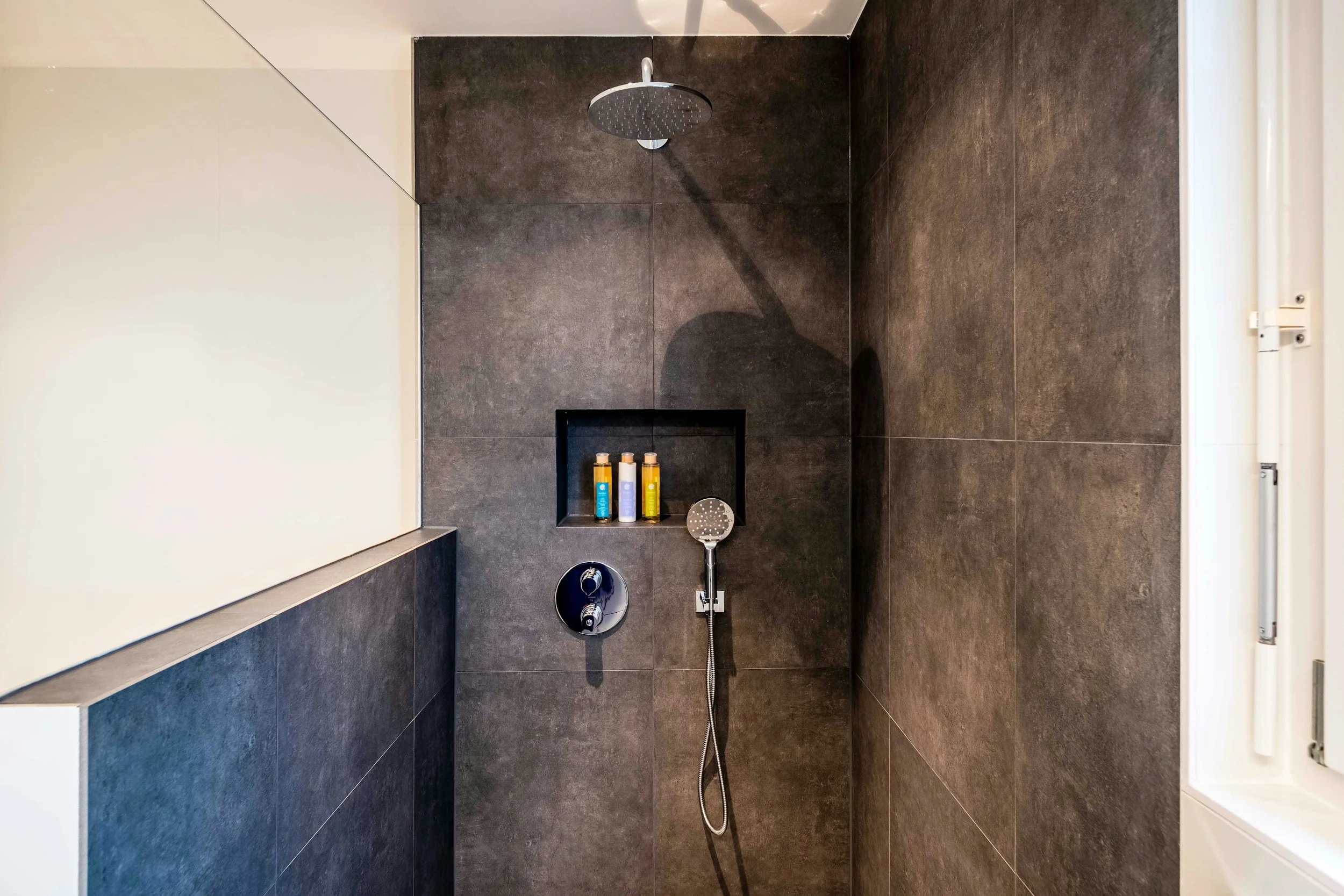 Modern shower area with large dark gray tiles, a round rainfall showerhead, a built-in shelf with toiletries, and a handheld shower on a hose.