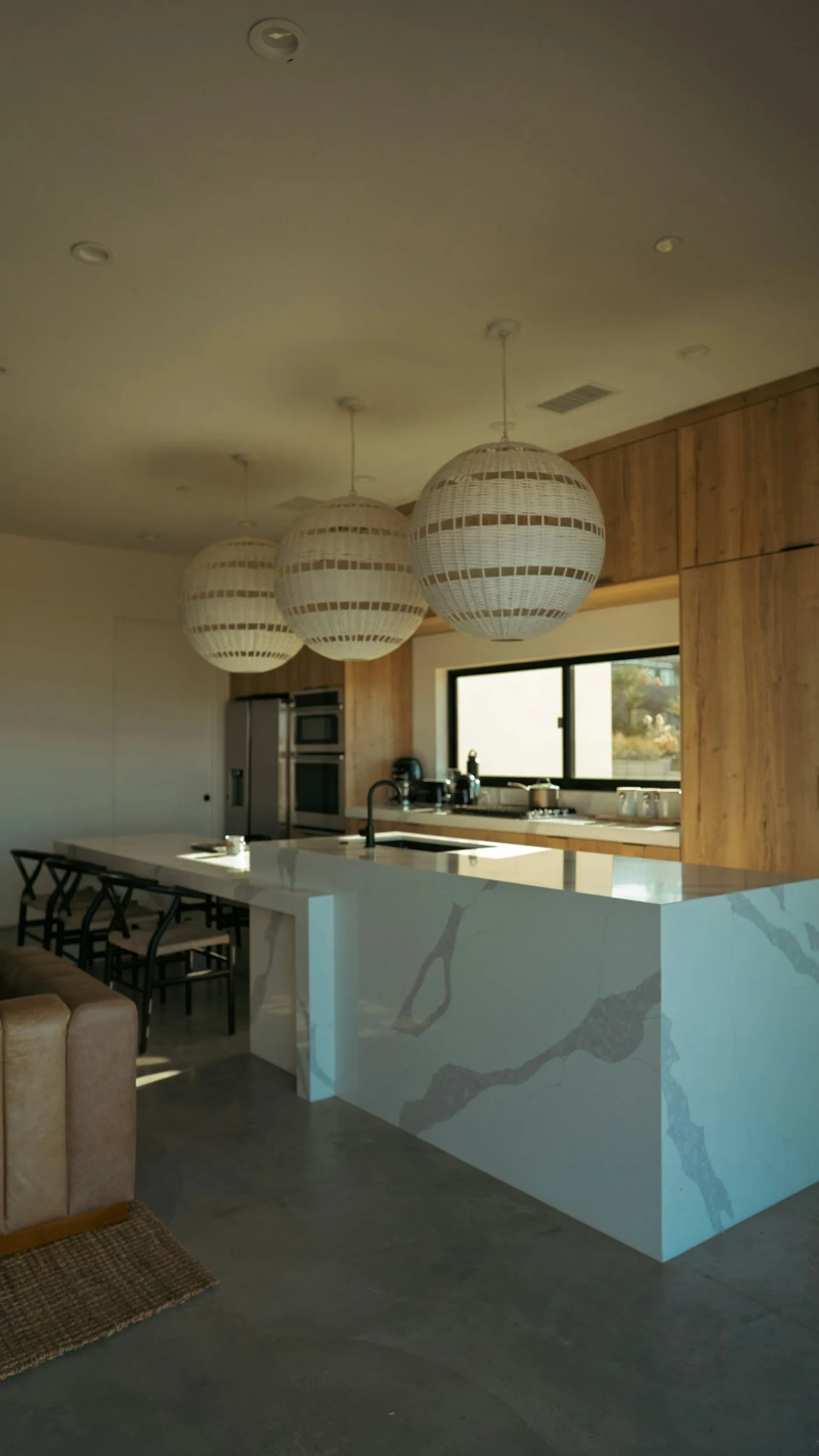 Modern kitchen with wooden cabinets, white marble island, black chandelier, and three large white woven pendant lights hanging from the ceiling, near a window with a view outside.