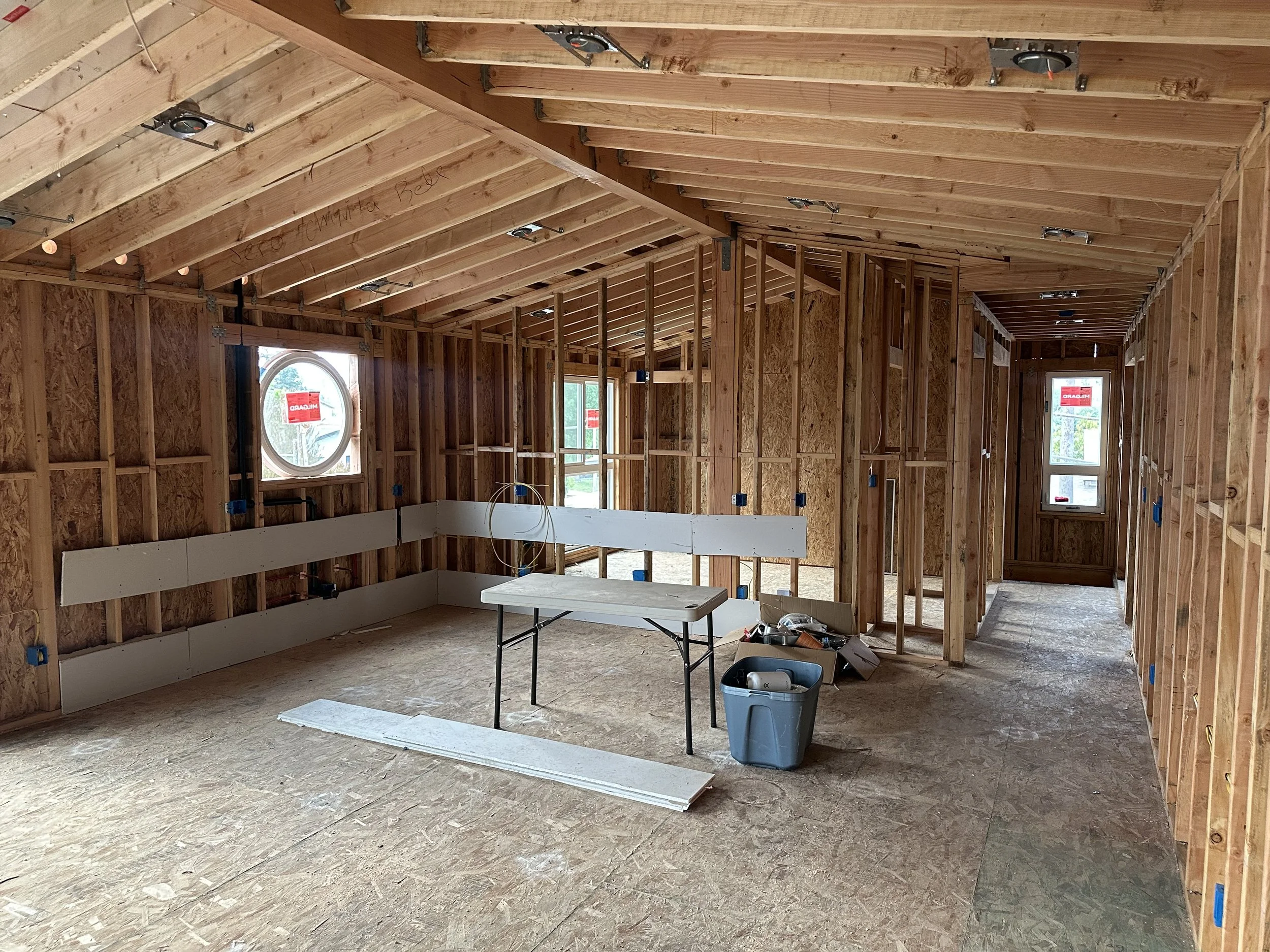Interior view of a house under construction with exposed wooden framing, multiple windows, electrical wiring, and a work table in the center.