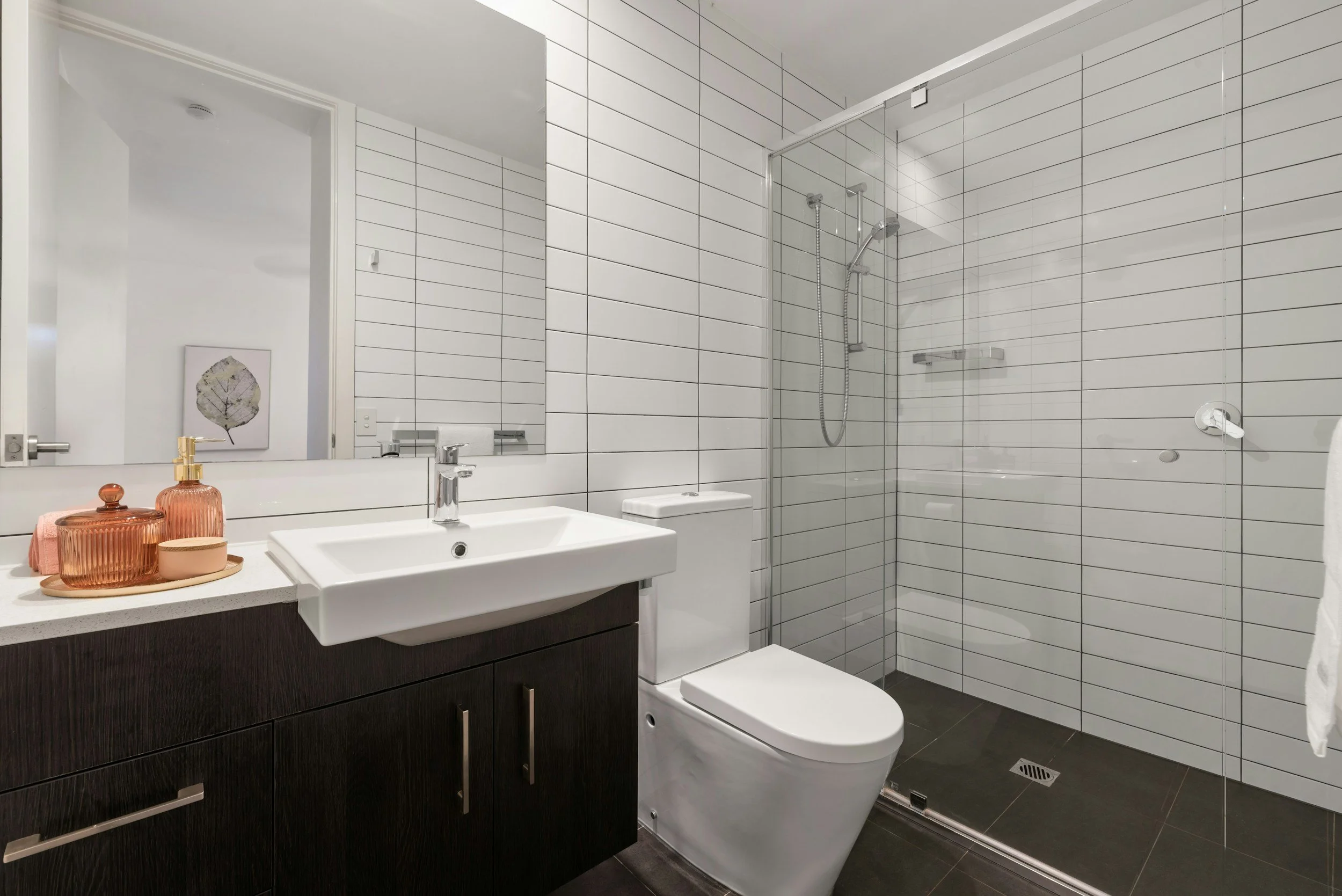 Modern bathroom with white tiled walls, a dark wood vanity with a white sink, toothbrush holders, a toilet, and a glass-enclosed shower area with a handheld showerhead and a drain on the dark tiled floor.