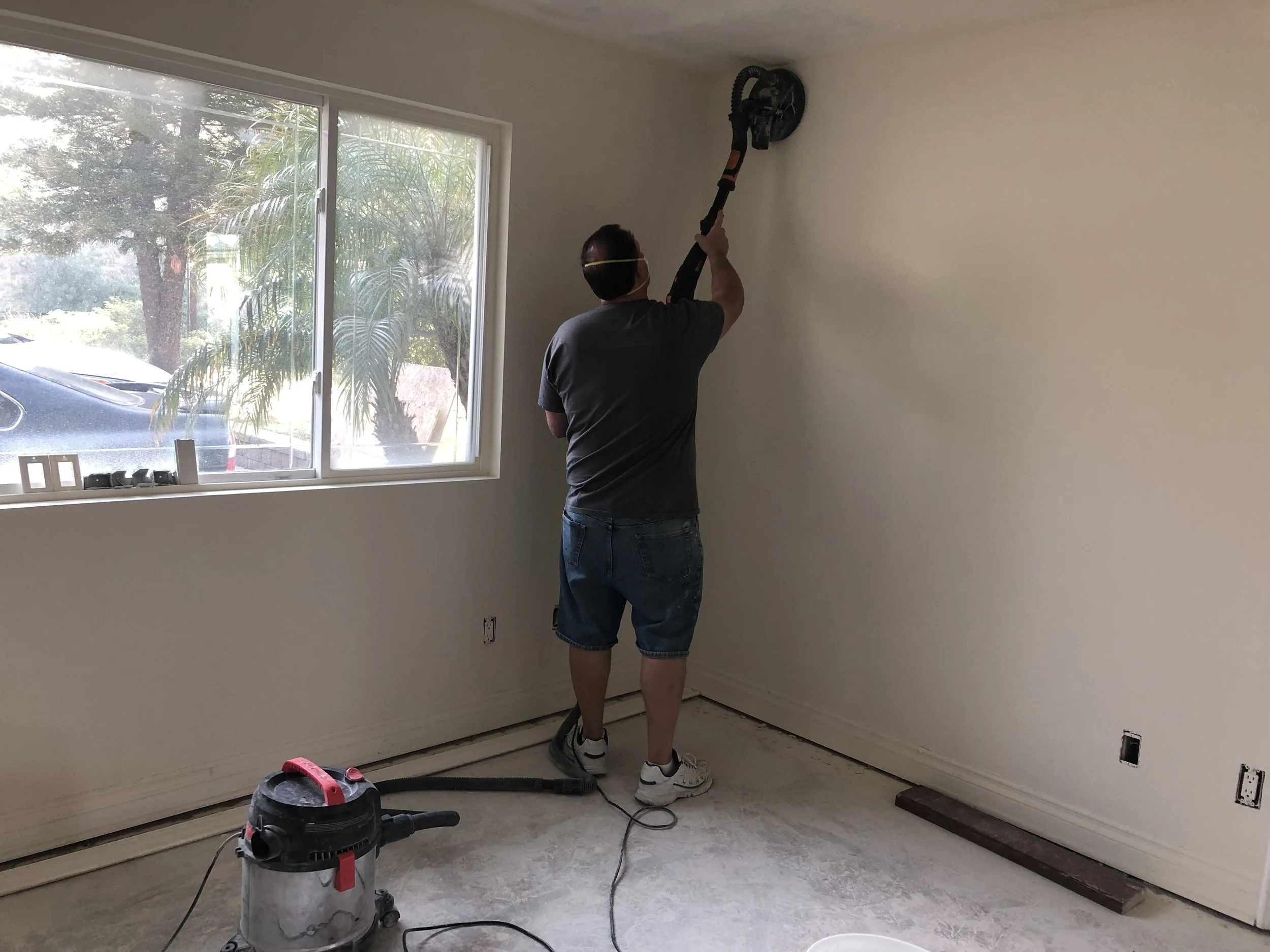 A person using a drywall sander on a ceiling in a room with a window and a vacuum cleaner on the floor.